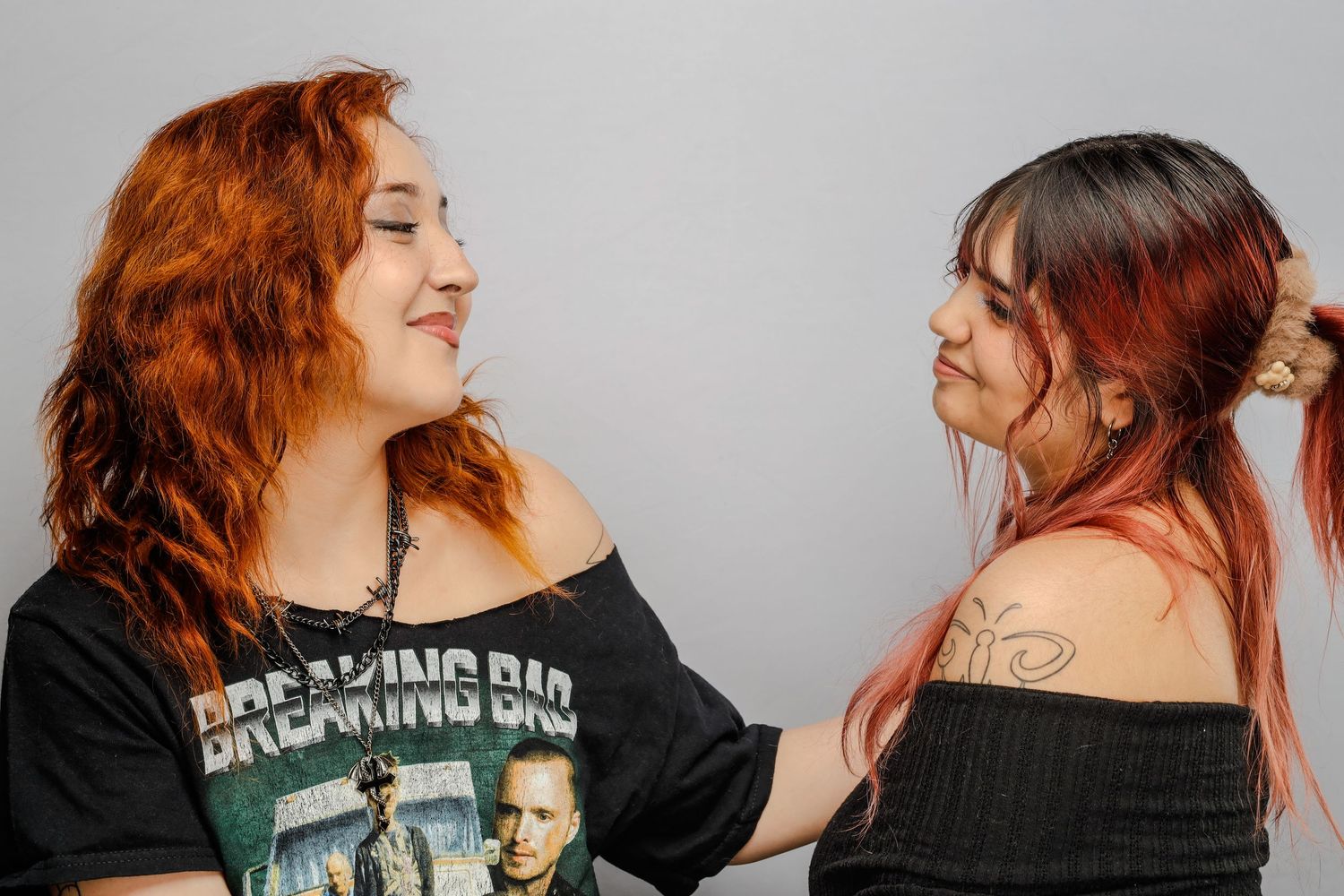 Two women with red hair wearing black tops share a candid laughing moment against white background.