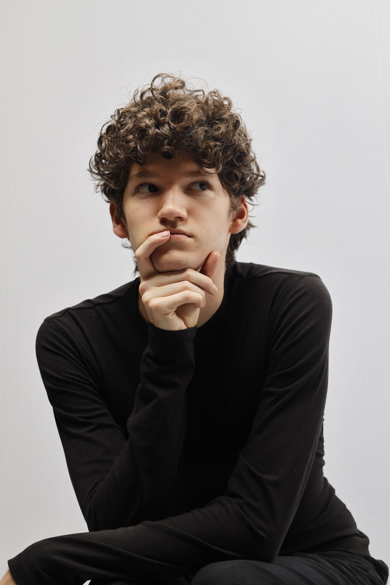Figure in a black sweater with curly hair sits contemplatively with hand on chin against white background.