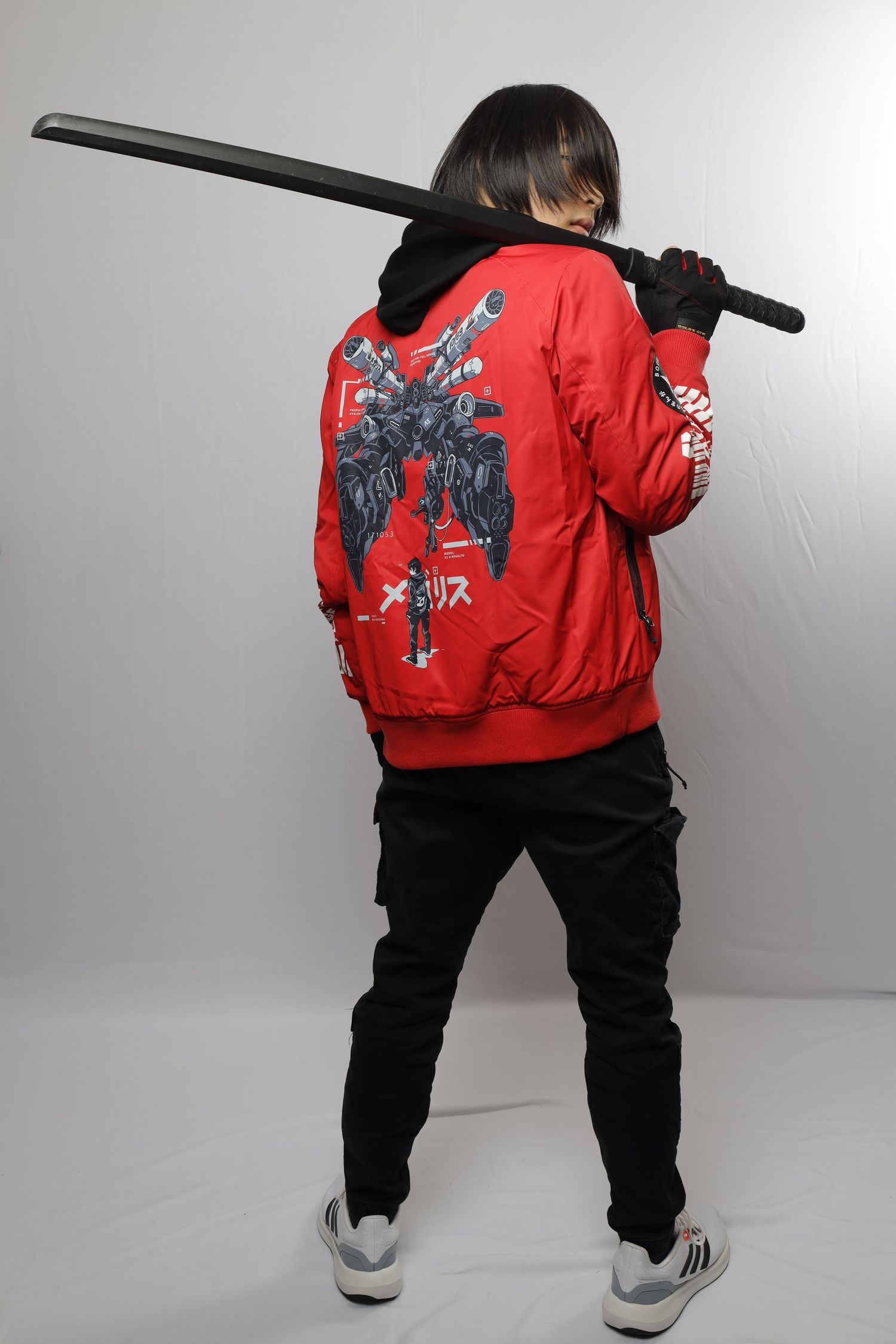 Person in red bomber jacket with graphic design on back poses holding a katana while wearing black pants and sneakers.
