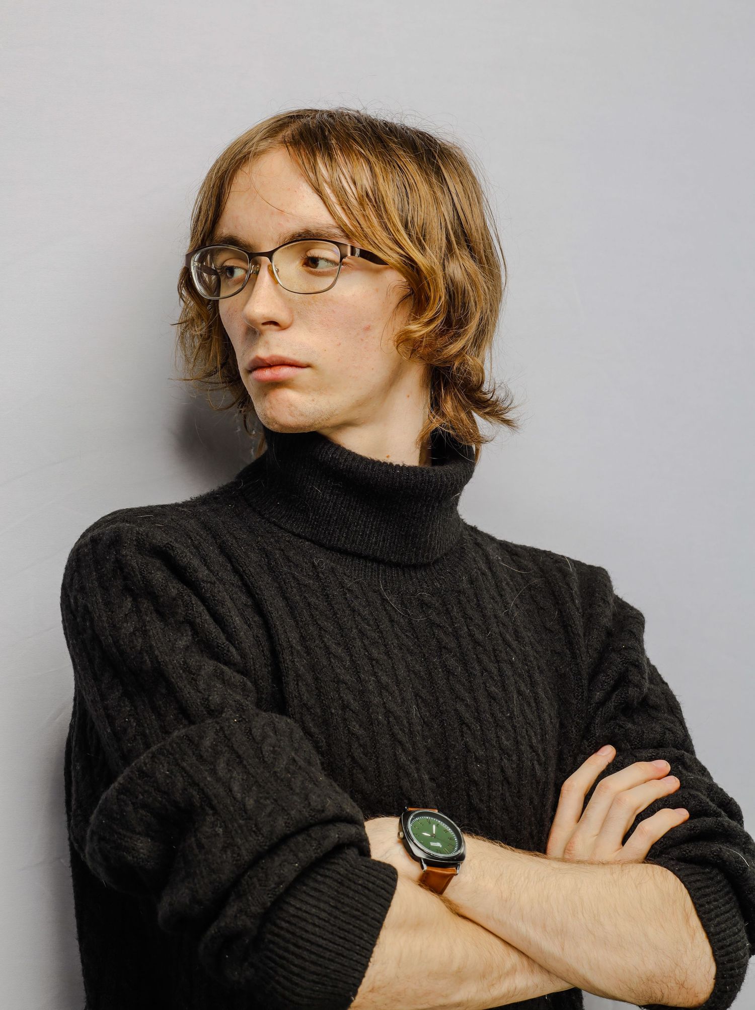Individual in black turtleneck sweater and glasses poses with crossed arms against gray background.
