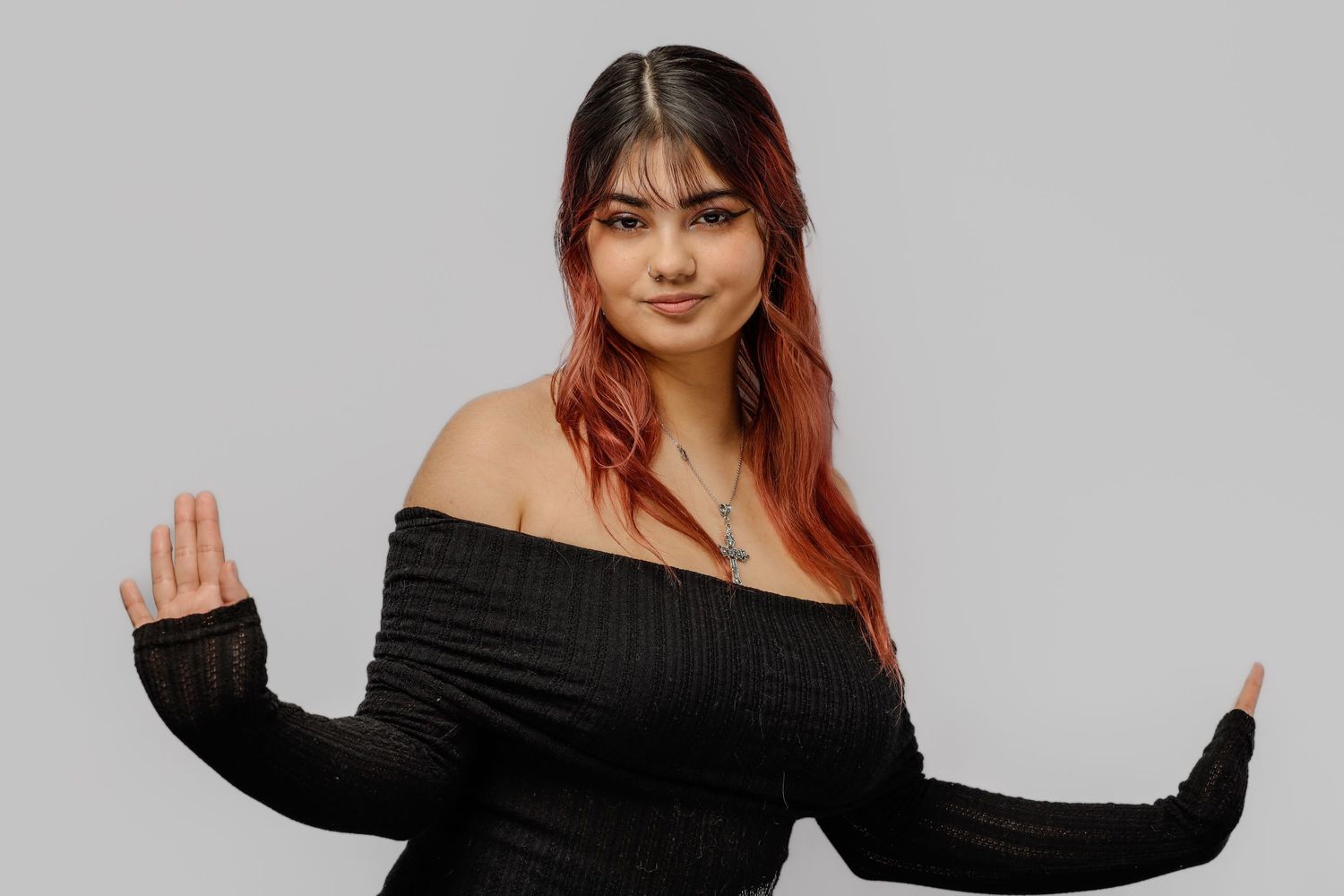 Person with reddish hair wearing a black off-shoulder sweater poses with arms outstretched against grey background.