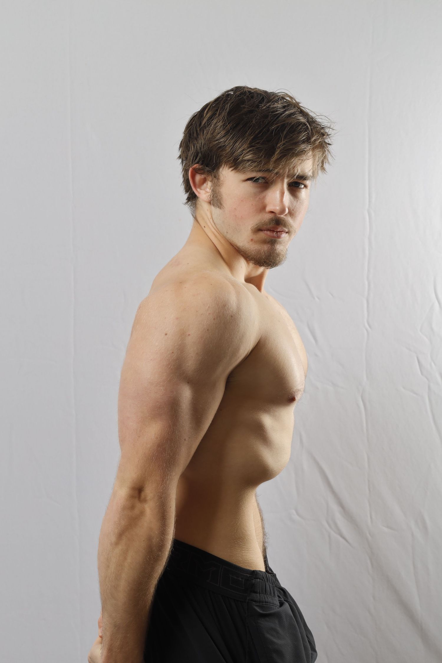 Athletic shirtless person with tousled hair wearing black shorts poses against white backdrop, showing muscular physique.