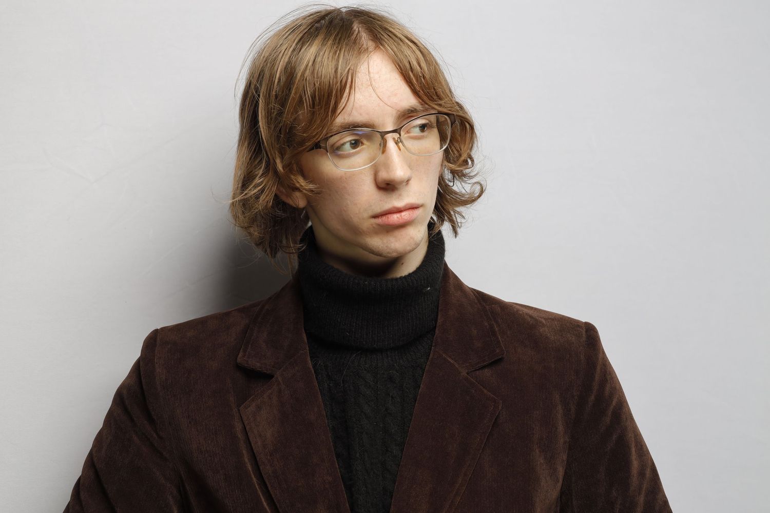 Studio portrait of someone in glasses wearing a brown corduroy jacket and black turtleneck.