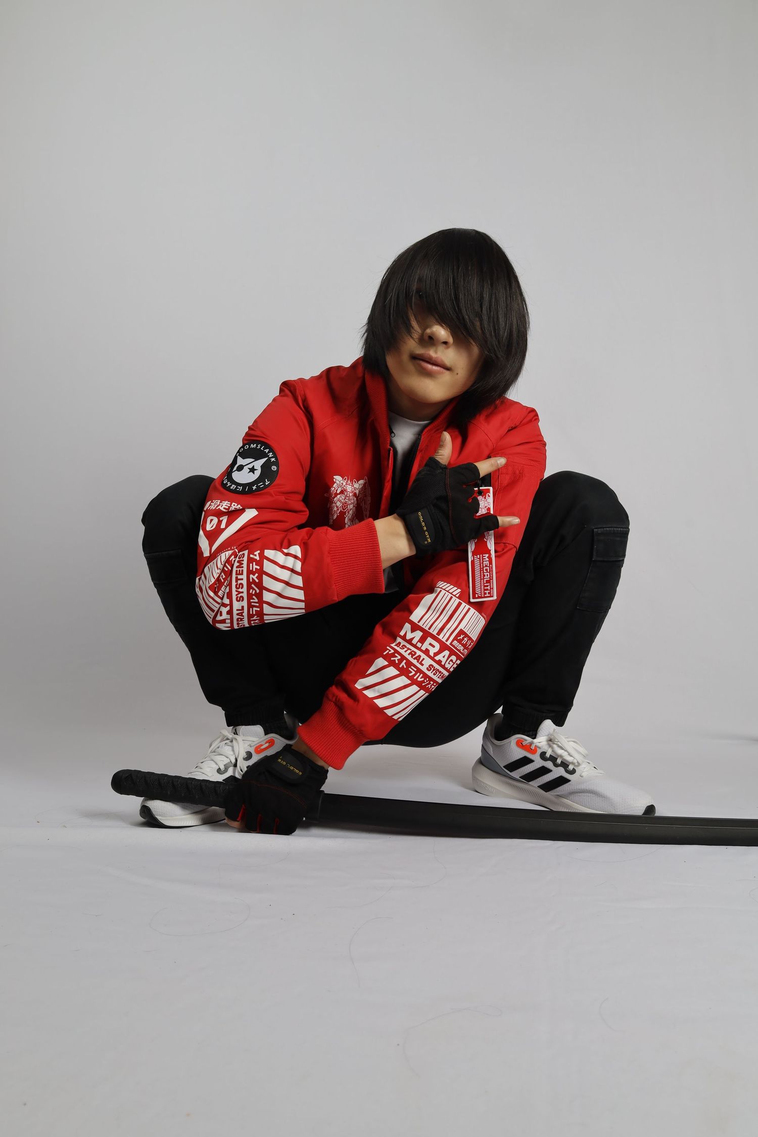 Fashion model in red Nike jacket and black pants strikes crouching pose against white backdrop wearing Adidas sneakers.