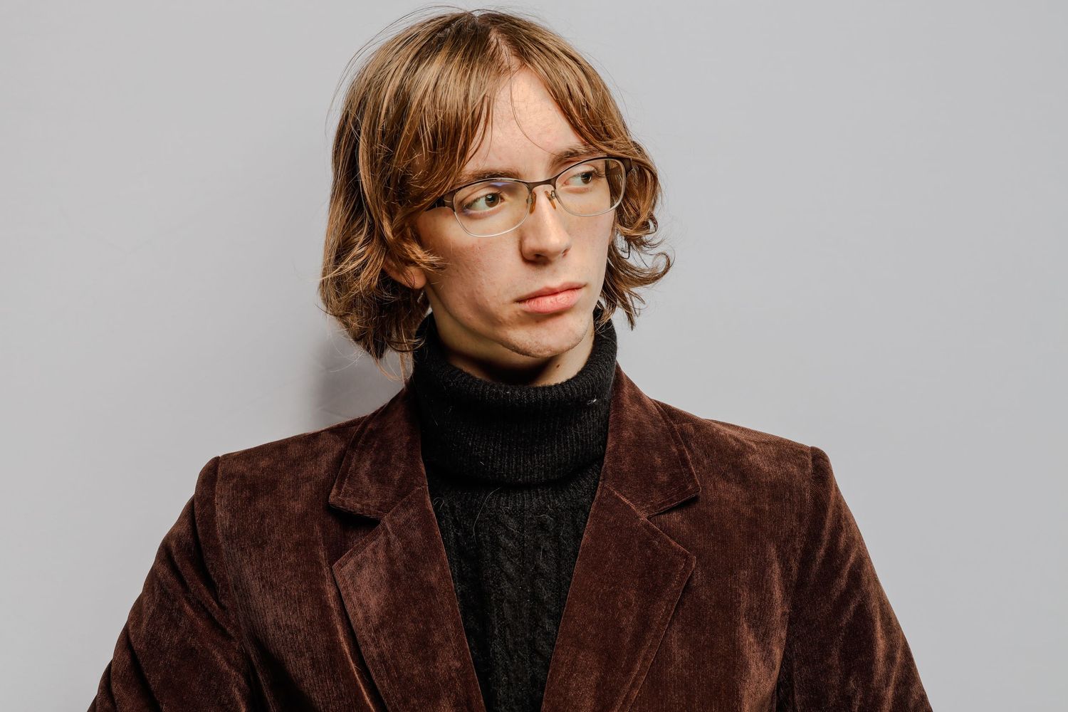 A person in rectangular glasses and a brown corduroy blazer over a black turtleneck sweater poses against a grey backdrop.