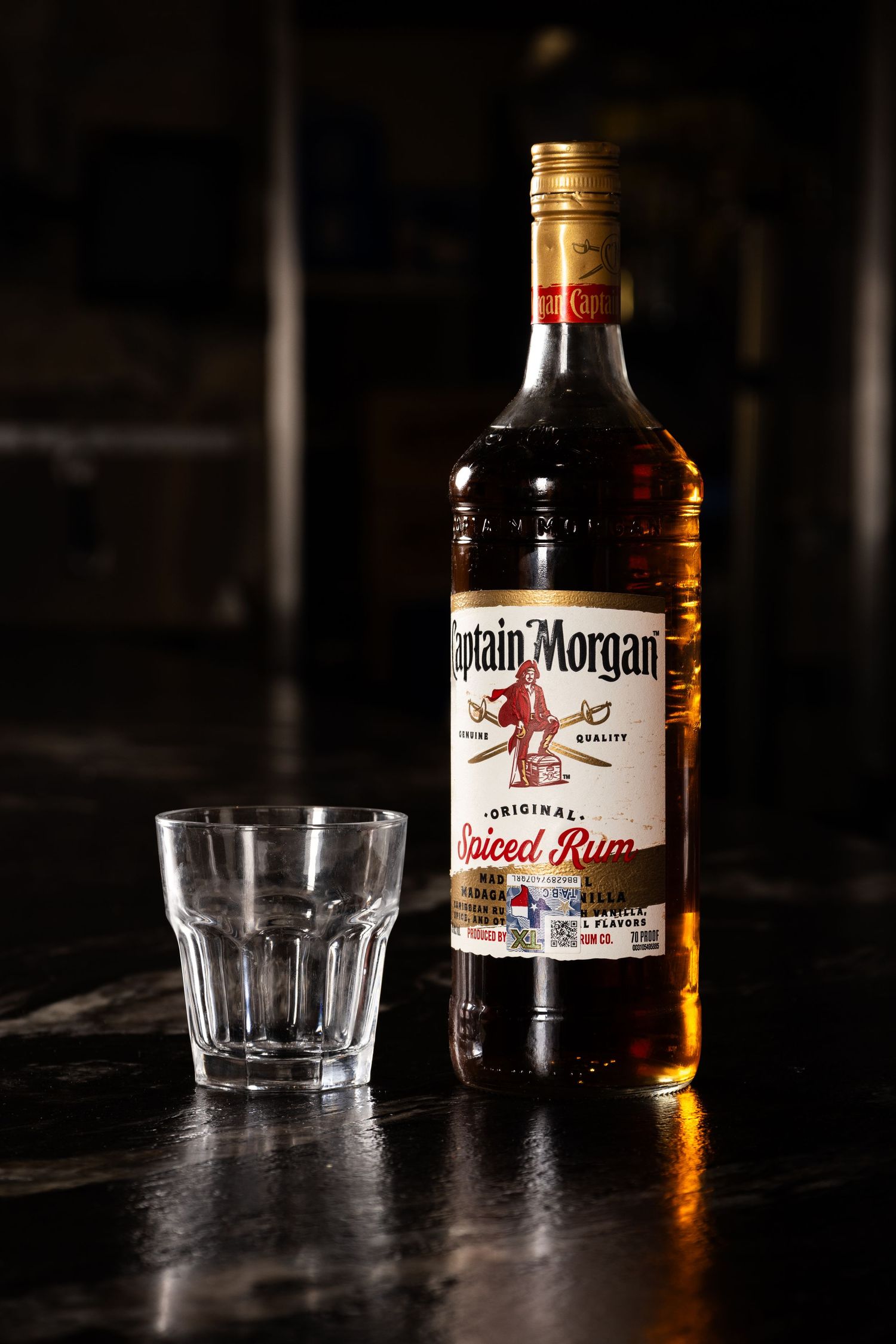 Bottle of Captain Morgan spiced rum with empty glass on dark wooden surface.