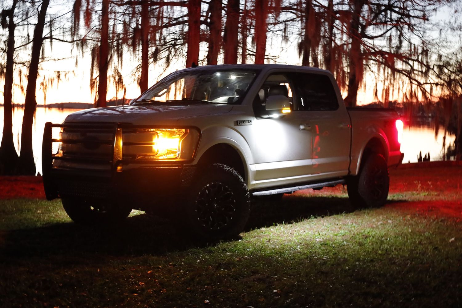 A silver Ford F150 pickup truck with illuminated headlights and tail lights parked at dusk under moss-covered trees.