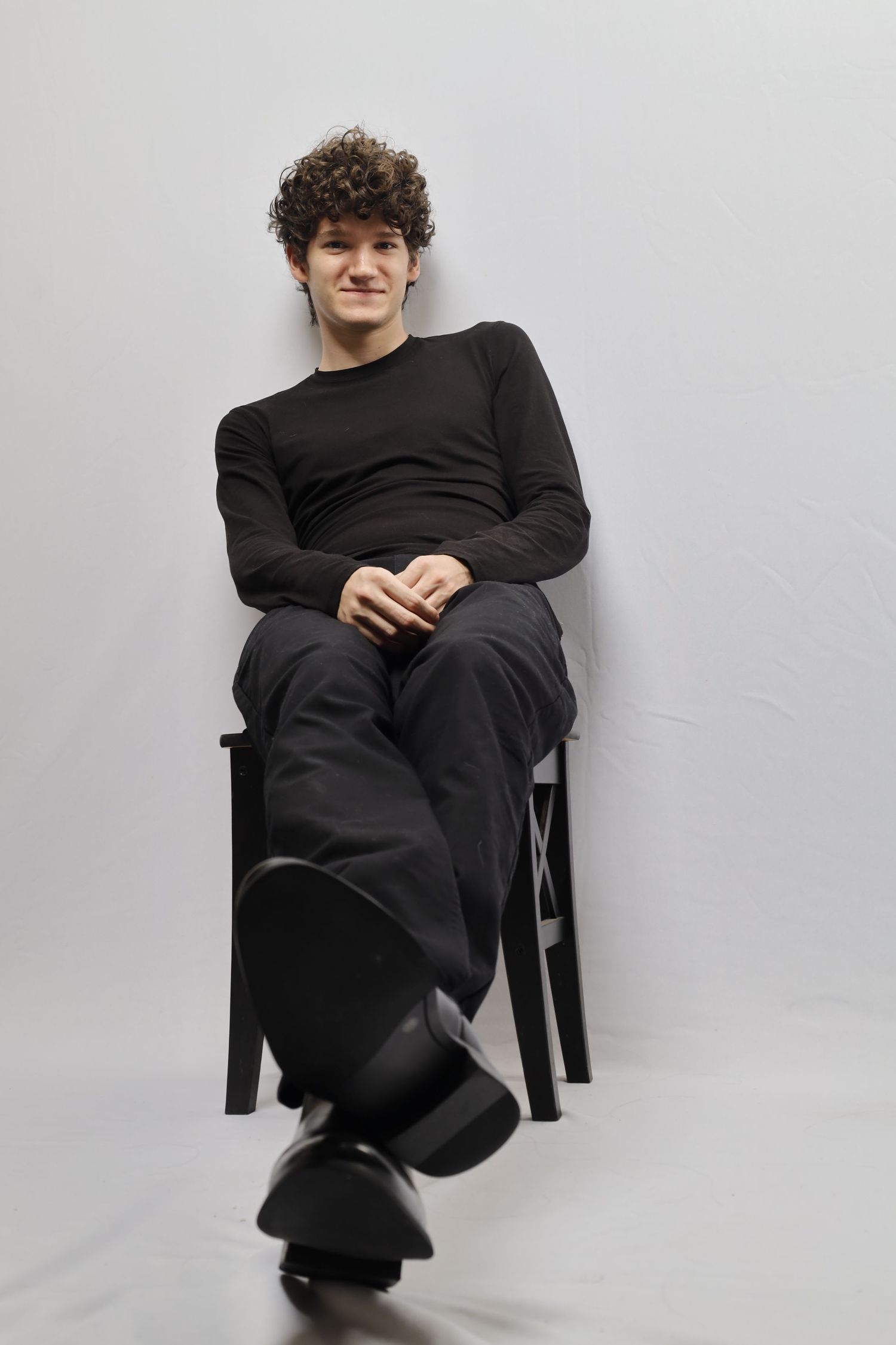 A person with curly dark hair wearing all black casual attire sits relaxed in a chair against a light gray background.