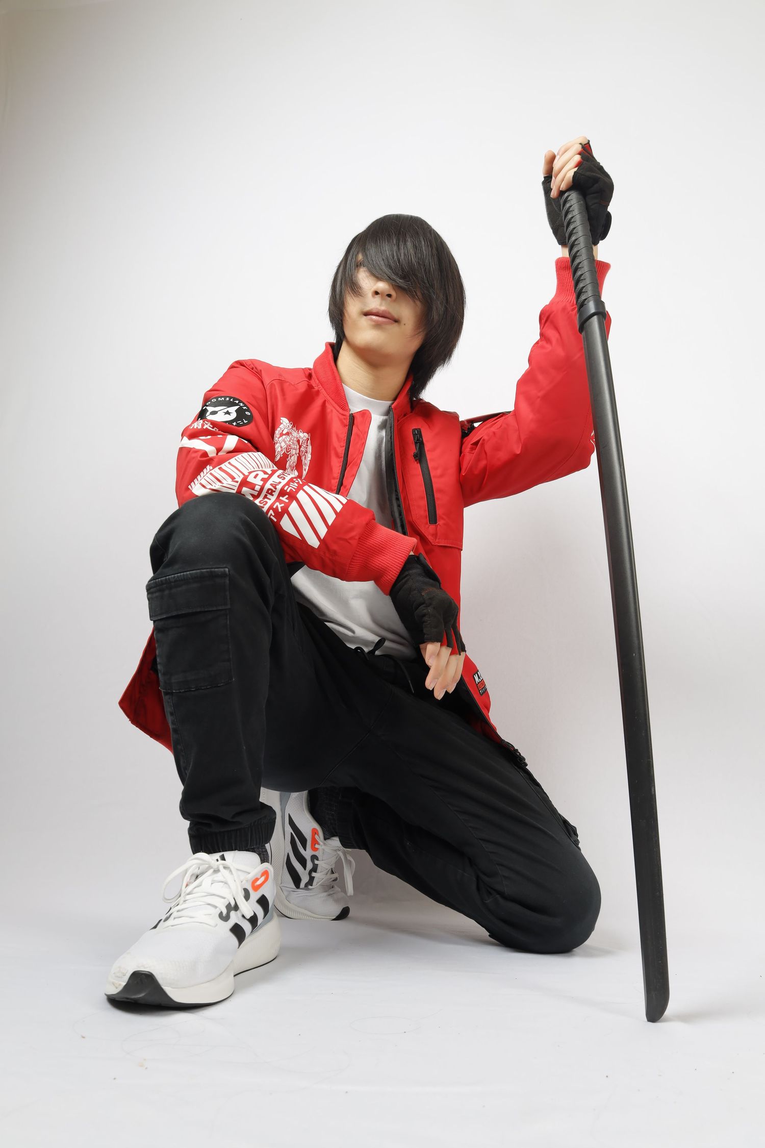 A person in a red jacket and black pants crouches while holding a long staff against a white background.