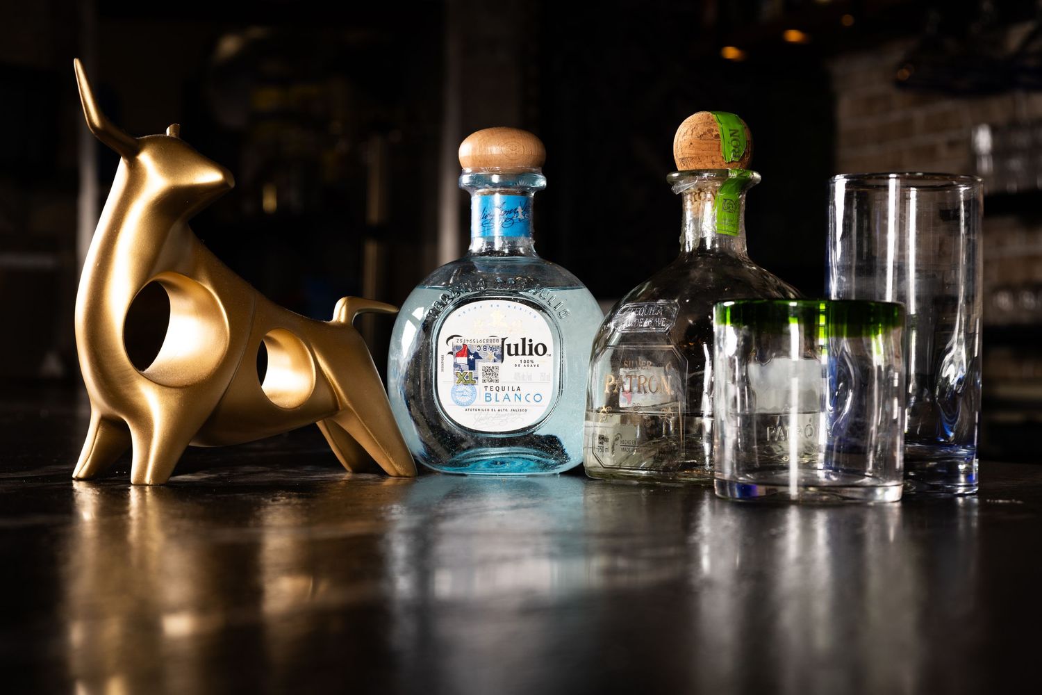 Various tequila bottles and glassware arranged on dark counter with decorative gold cat statue casting dramatic shadows.