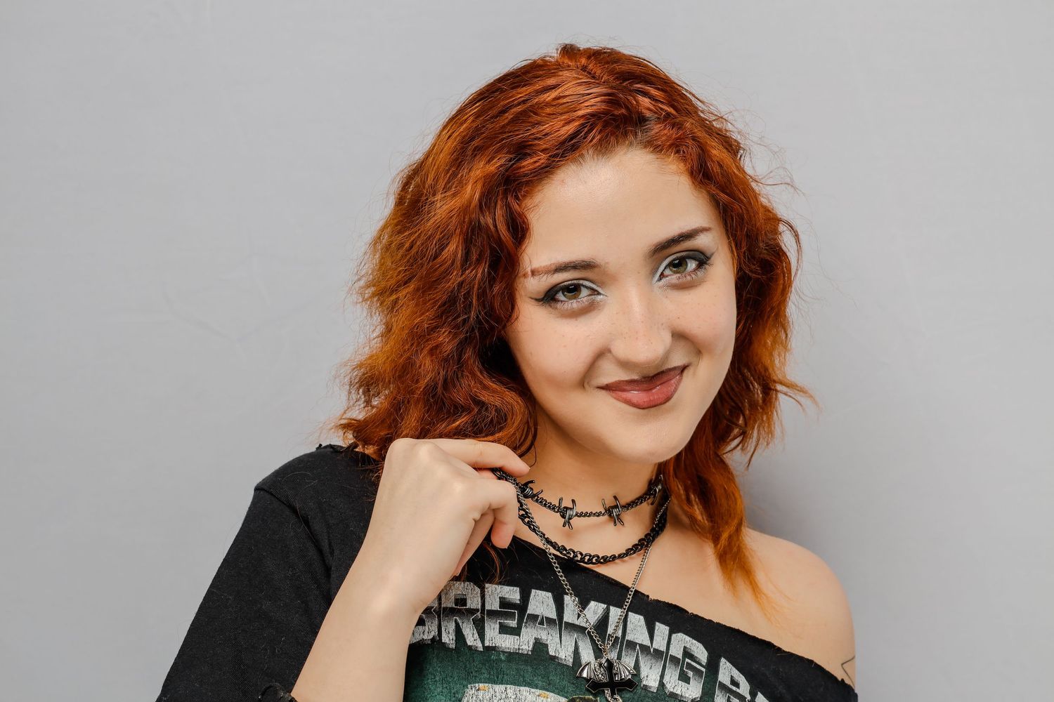Someone with vibrant curly red hair and a choker necklace smiles while wearing a black graphic t-shirt against a gray background.