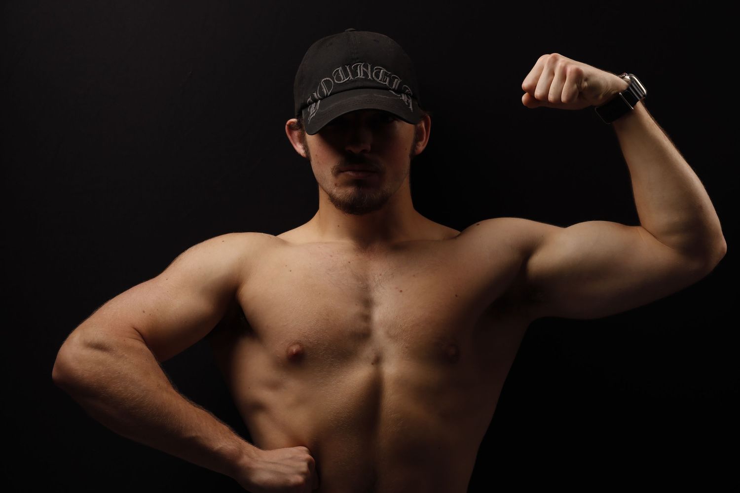 Shirtless athletic figure flexing bicep muscles while wearing a baseball cap against dark background.