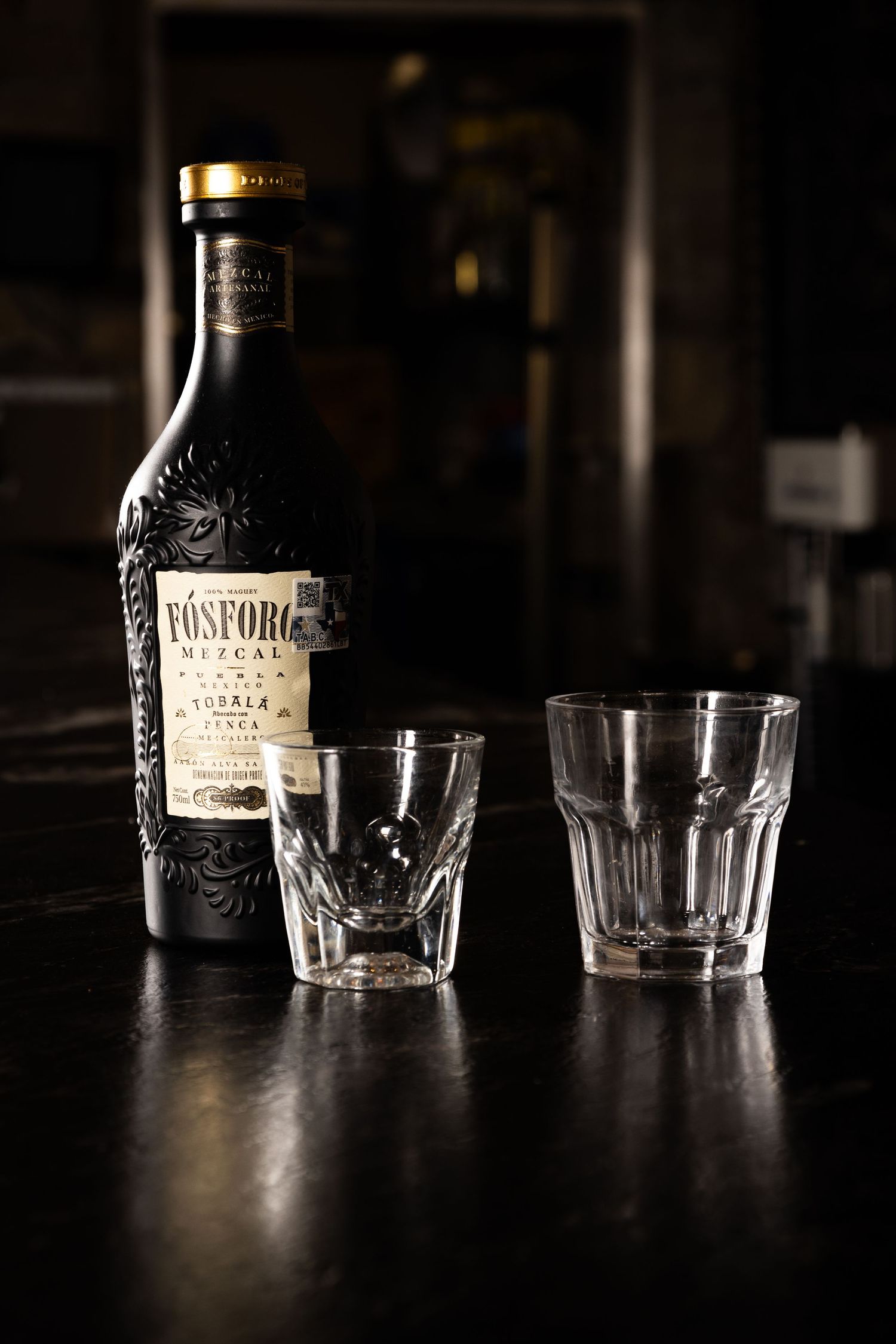 A dark bottle of Kinsford vodka stands beside two empty glass tumblers on a wooden surface in moody lighting.