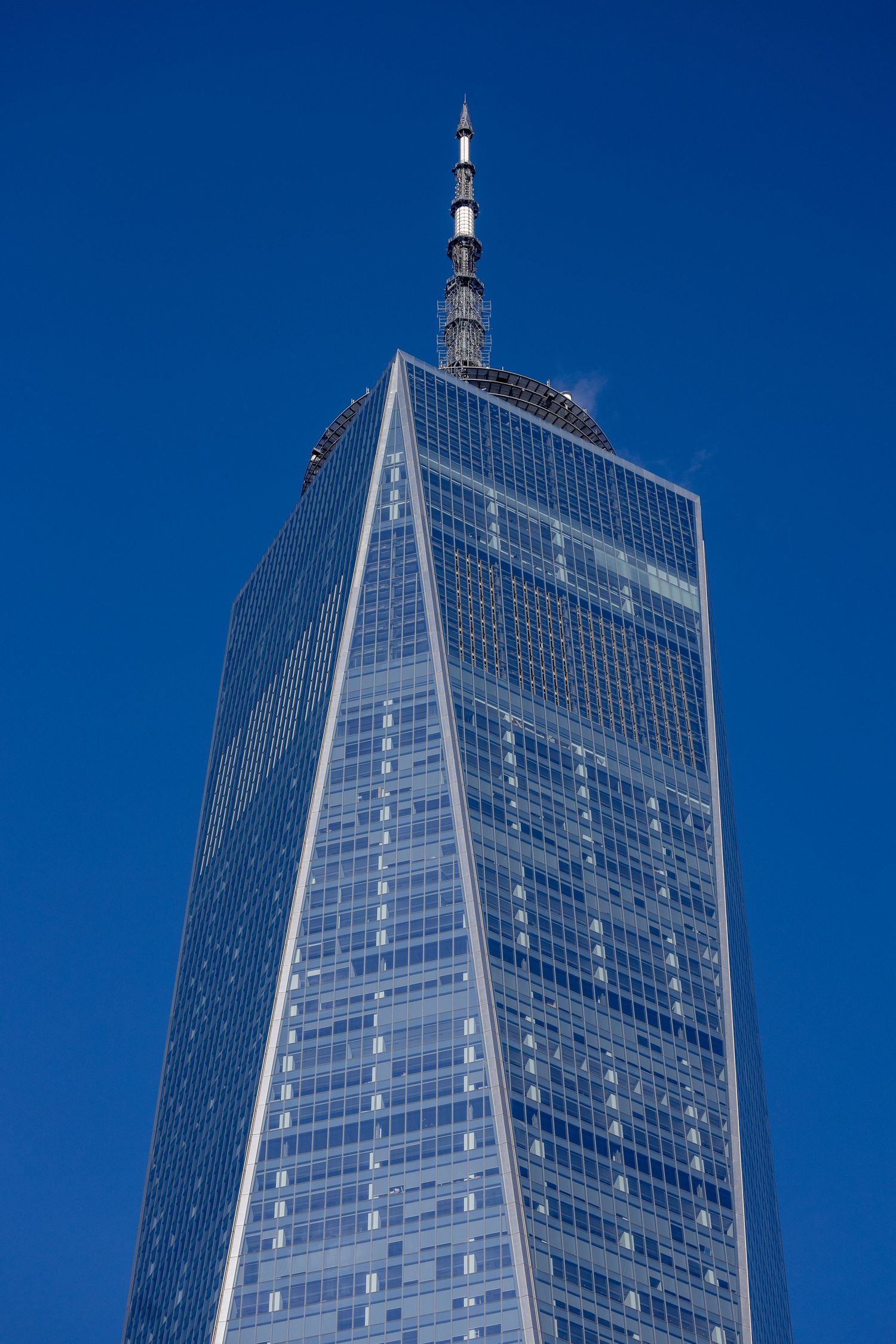 Looking up at One World Trade Center's gleaming glass exterior and spire against a deep blue sky in New York City.