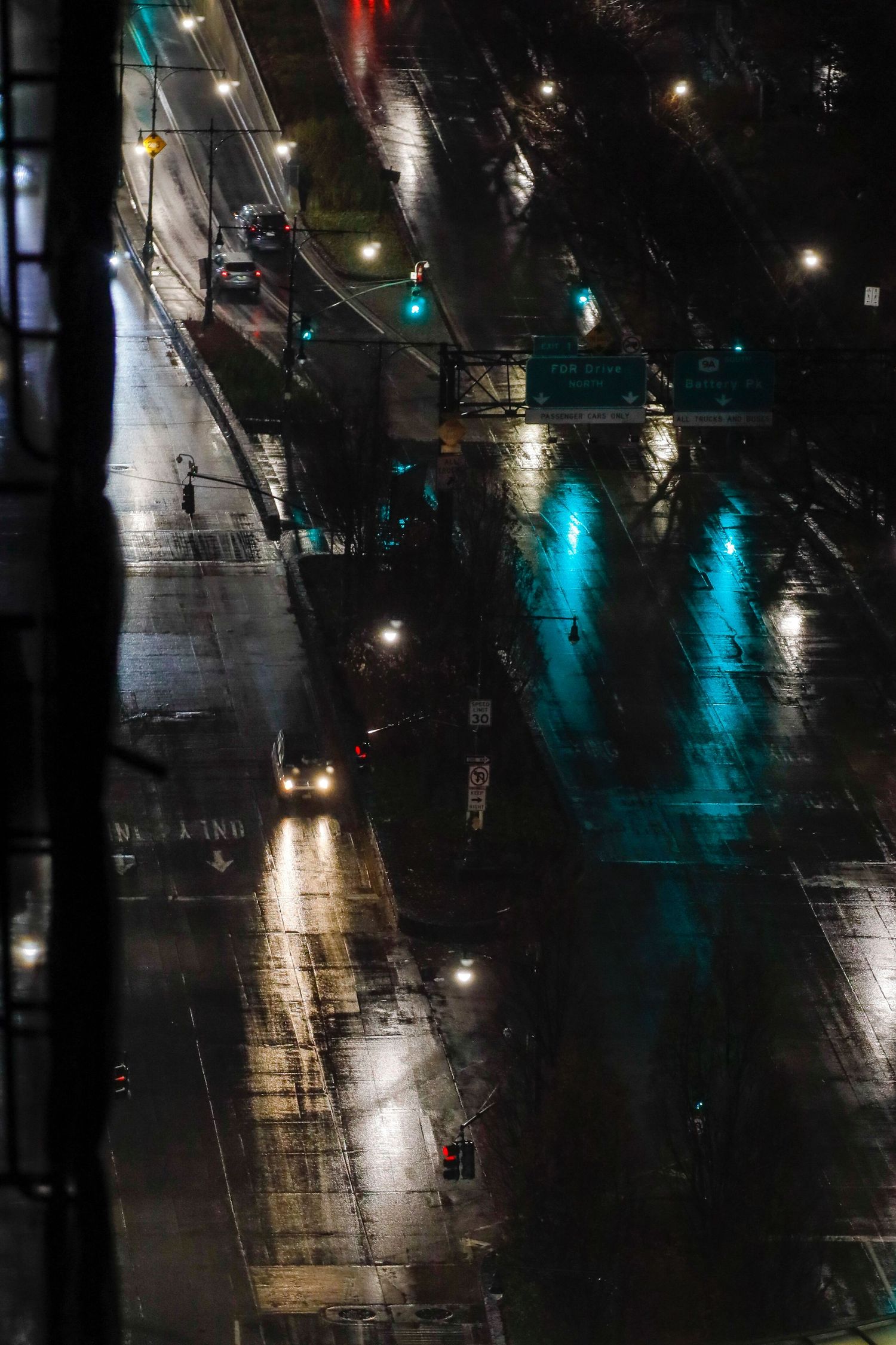 Aerial view of a rainy city street at night with reflective wet pavement and glowing traffic lights casting colorful reflections.