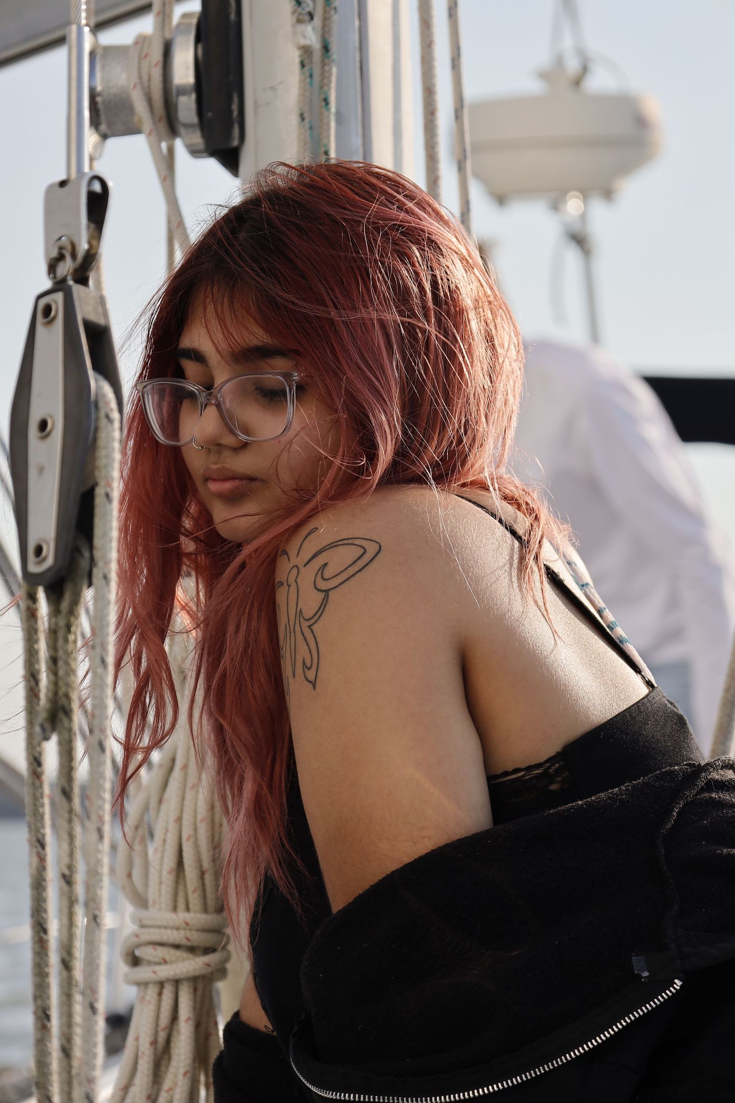 A red-haired person with glasses and shoulder tattoo leans against boat rigging.