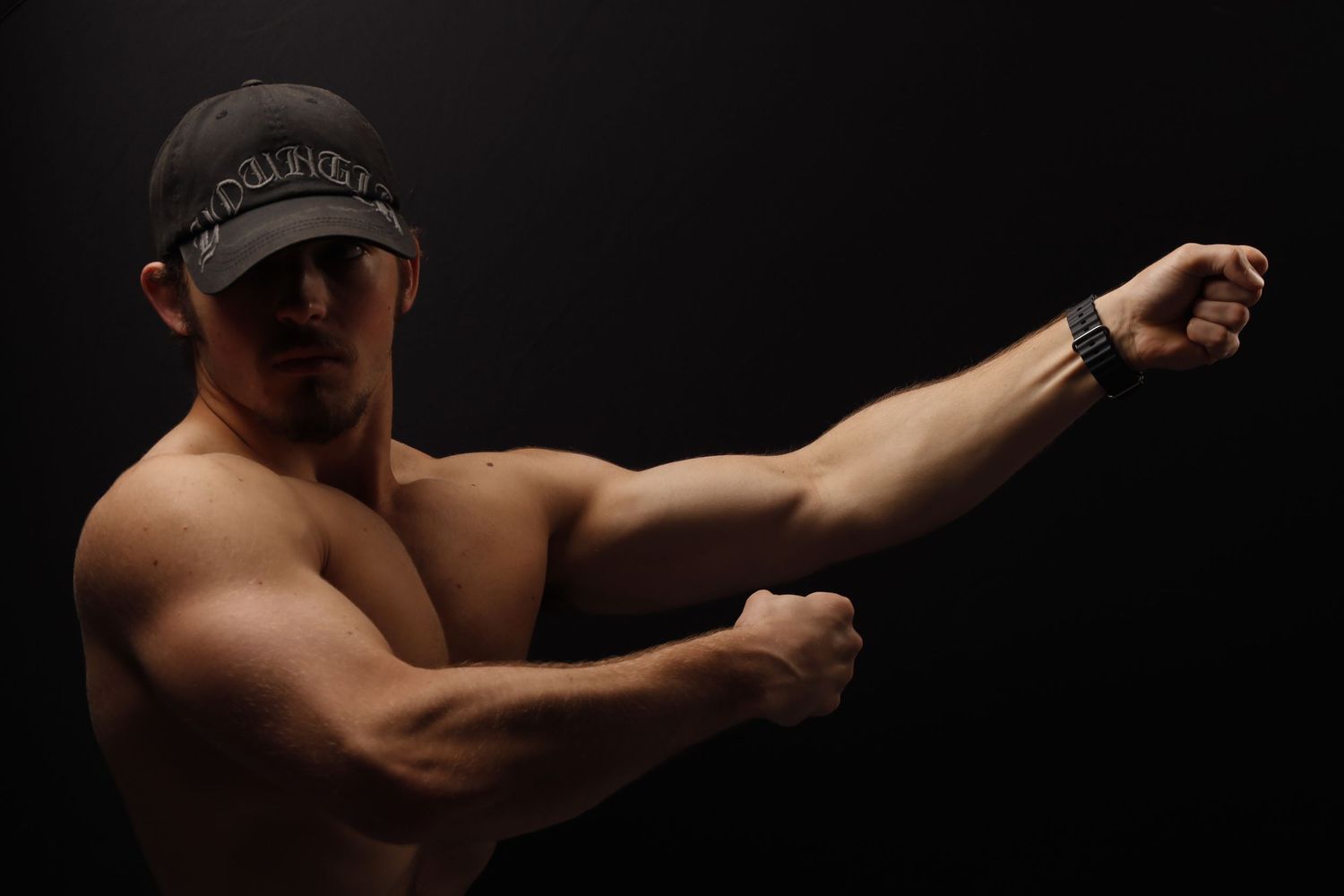 Muscular shirtless man wearing a black baseball cap strikes a dramatic boxing pose against dark background.