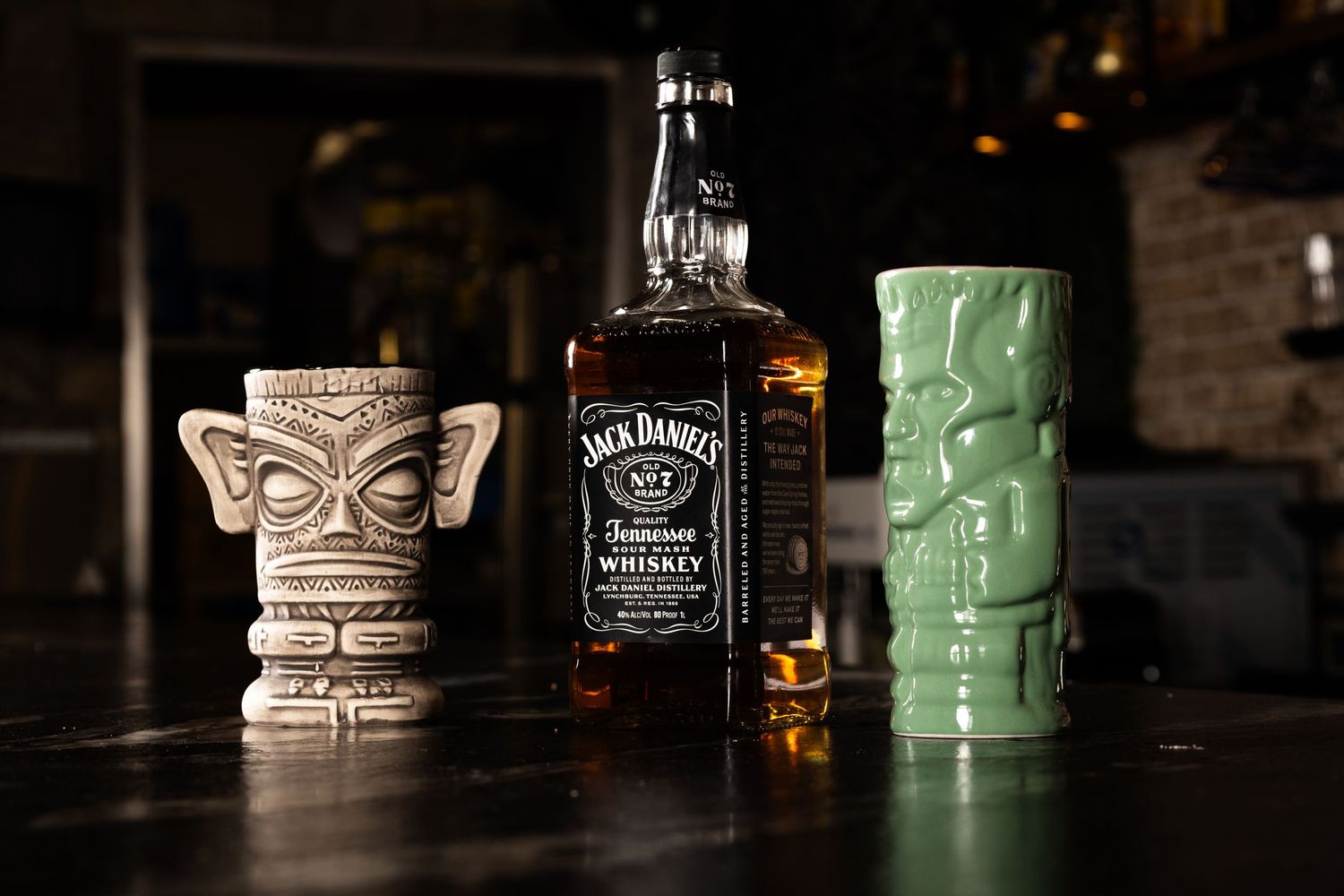 A collection of tiki mugs and a Jack Daniel's whiskey bottle arranged on a dark wooden bar surface.