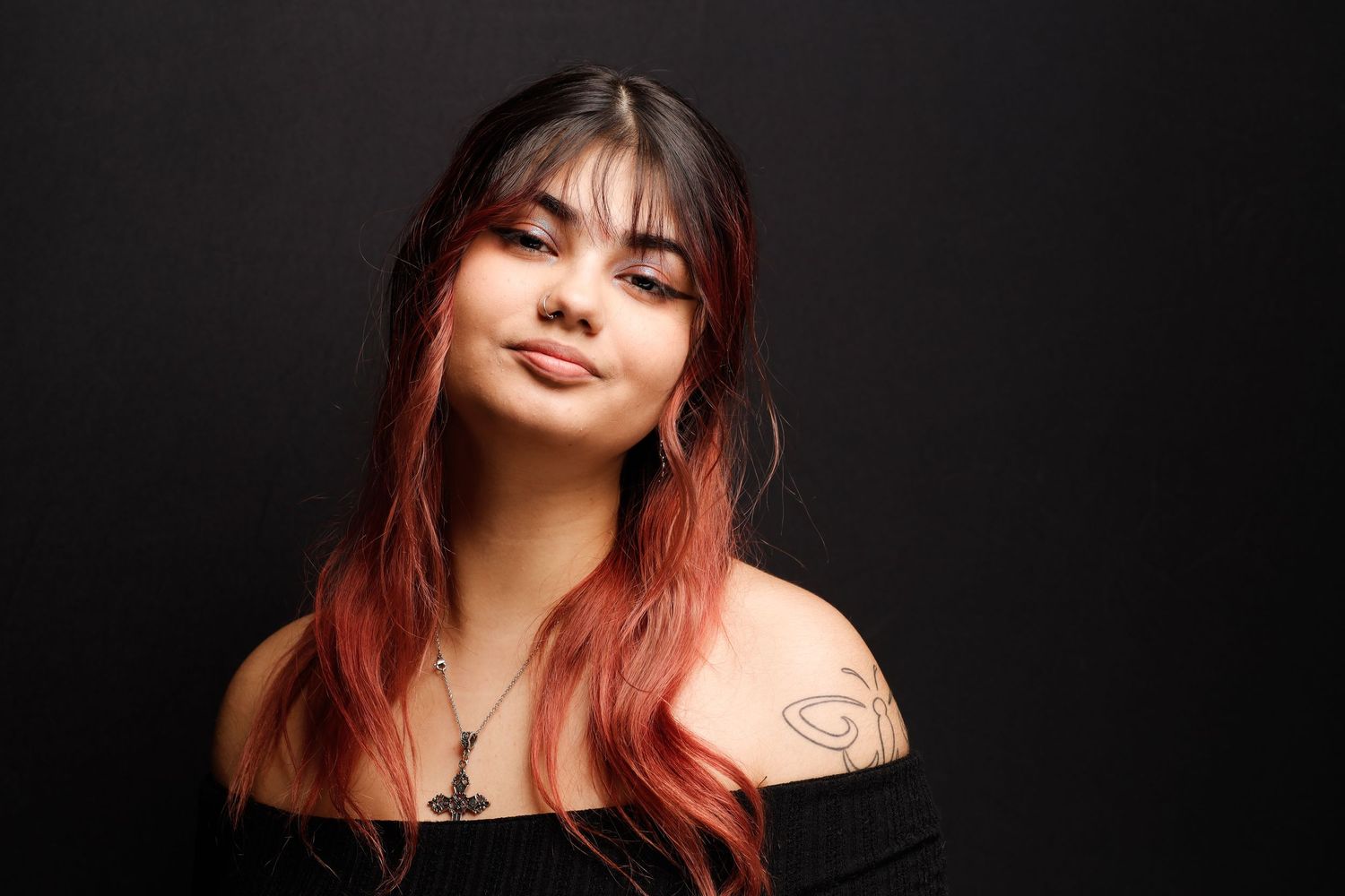 Portrait of someone with long black and pink ombre hair wearing a black off-shoulder top against dark background.