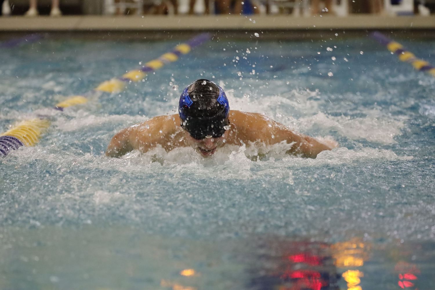 Competitive swimmer performing butterfly stroke in indoor pool with water splashing.