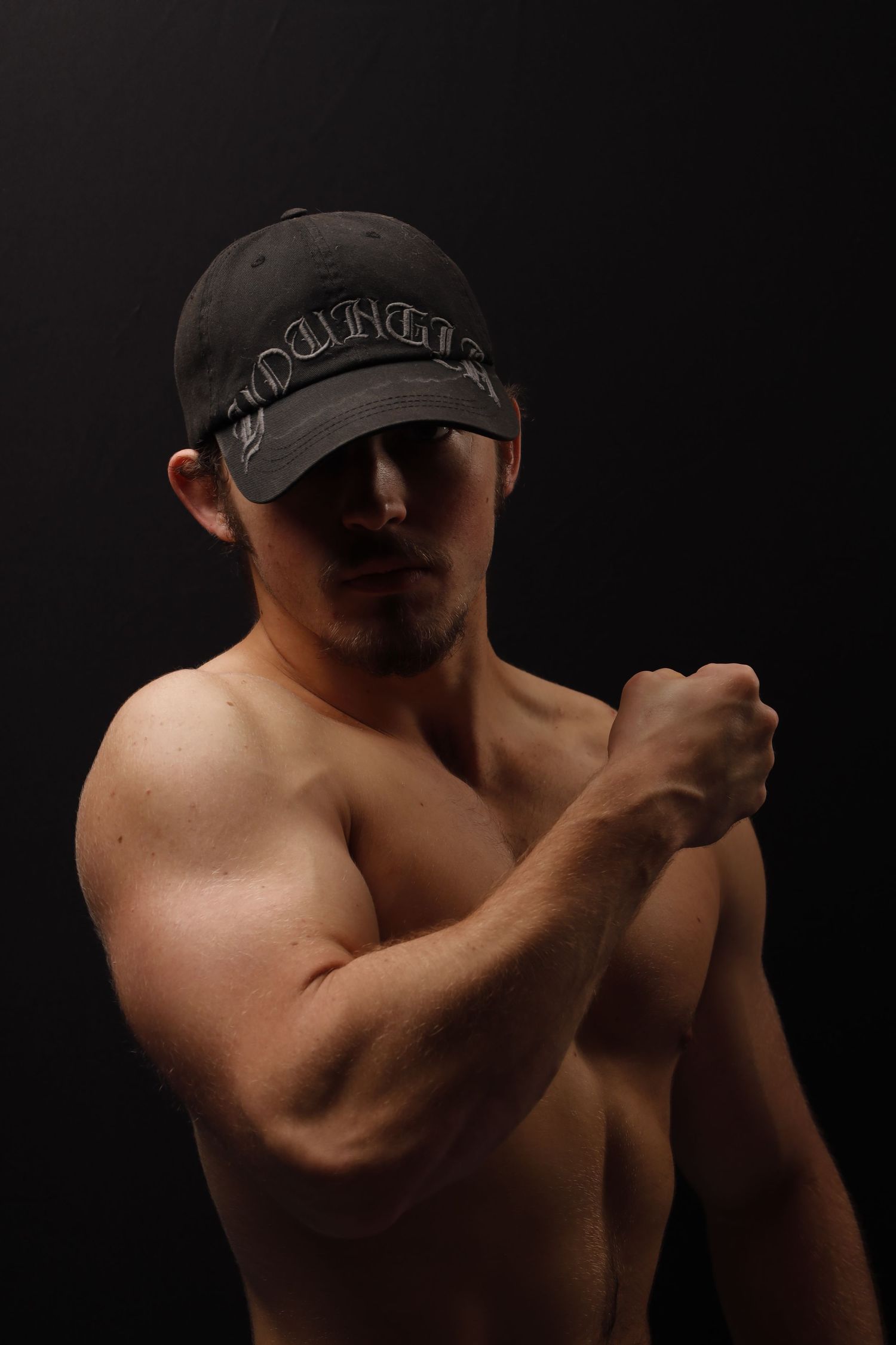 An athletic shirtless person in a dark baseball cap poses with raised fists against a black background.