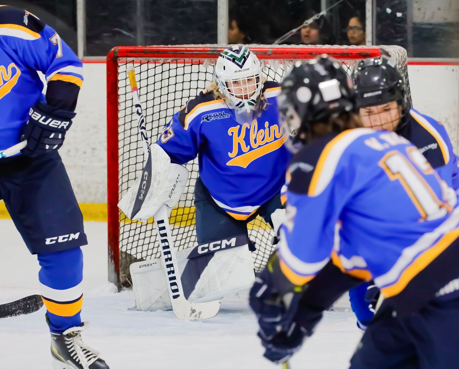 Ice hockey goalie in blue jersey guards net during intense game action with players nearby.