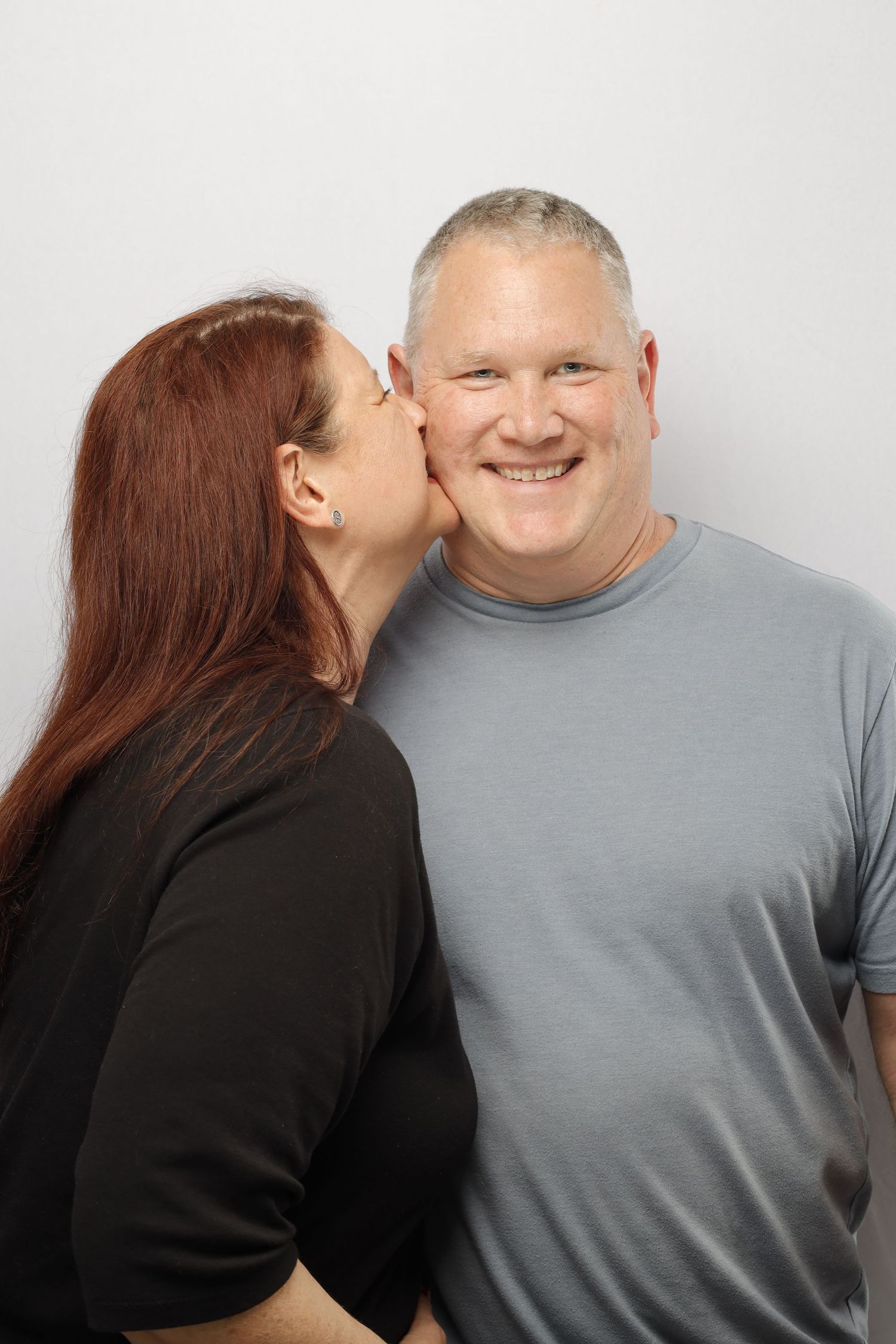 A woman with long red hair kisses the cheek of a person wearing a gray t-shirt against a white background.