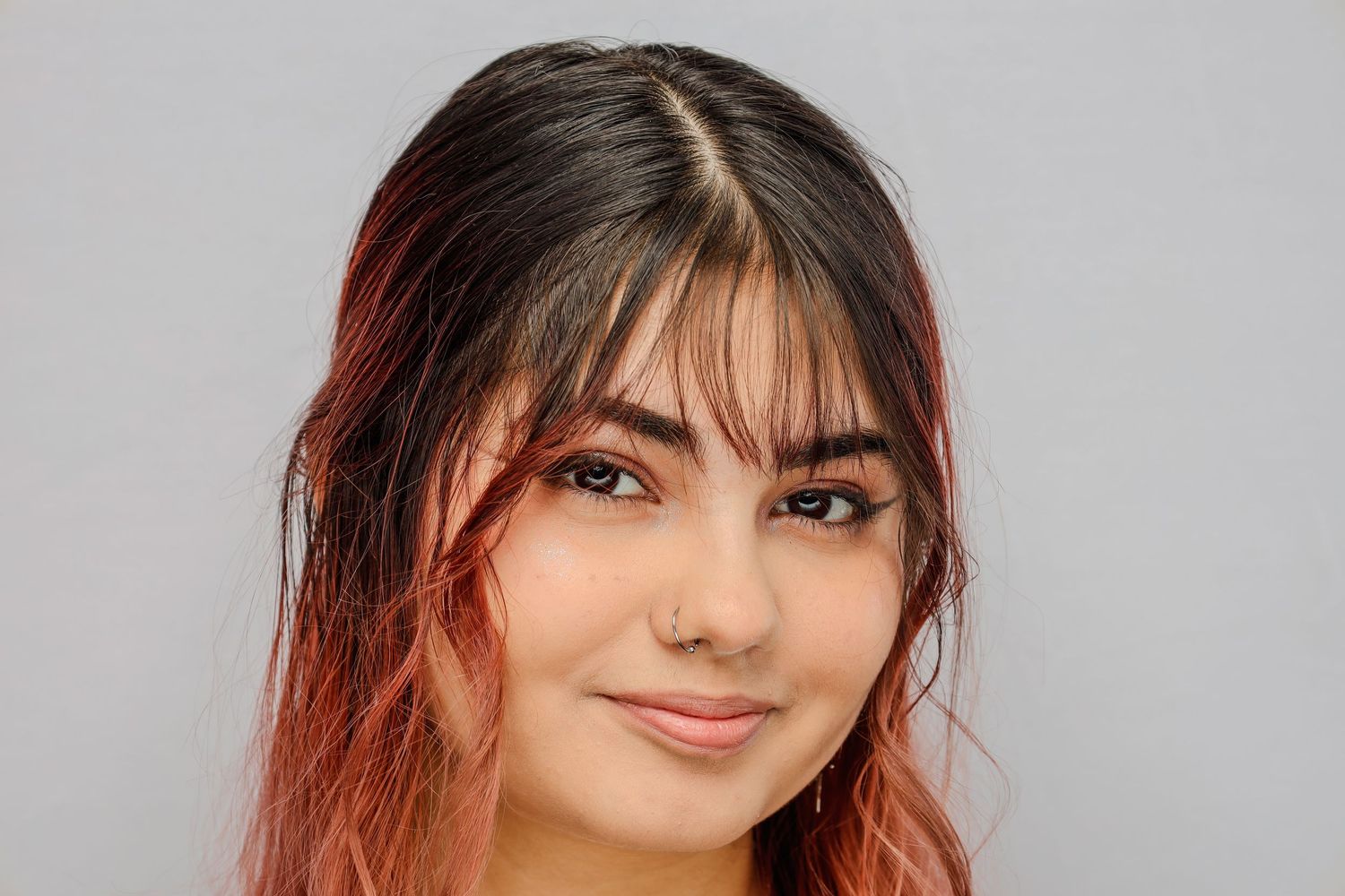 Portrait of a person with long dark hair with red highlights looking directly at camera against a neutral background.