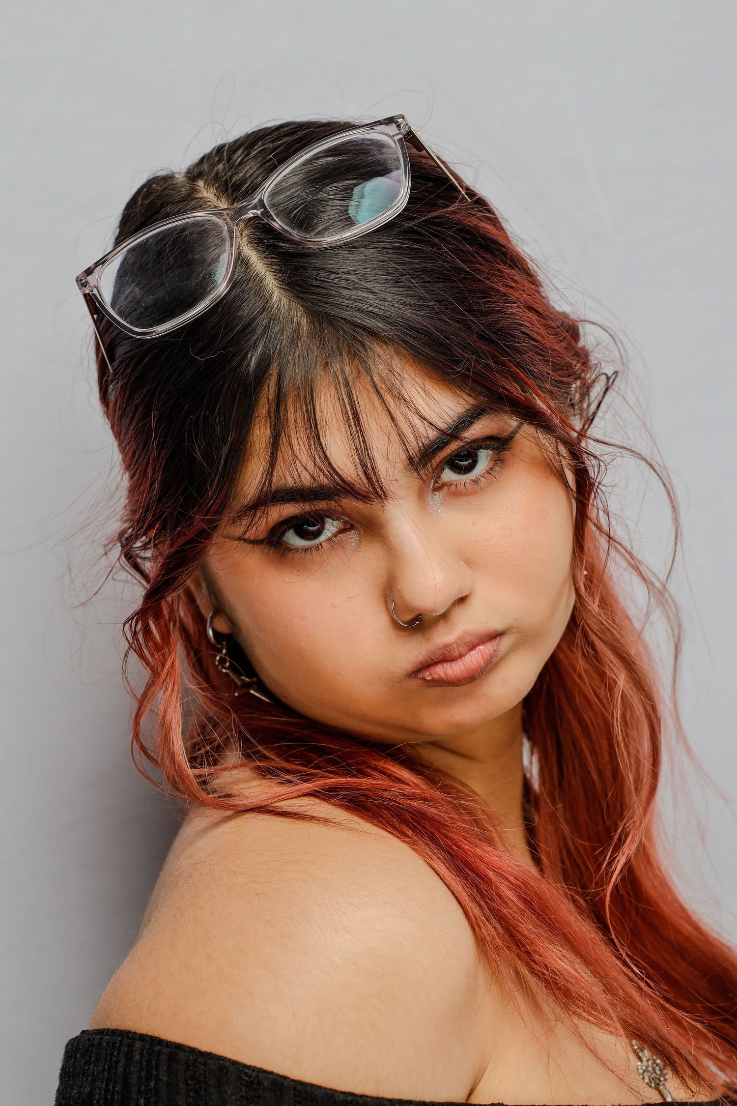 Close-up portrait with glasses perched on top of dark and red ombre hair, wearing an off-shoulder black top against gray backdrop.