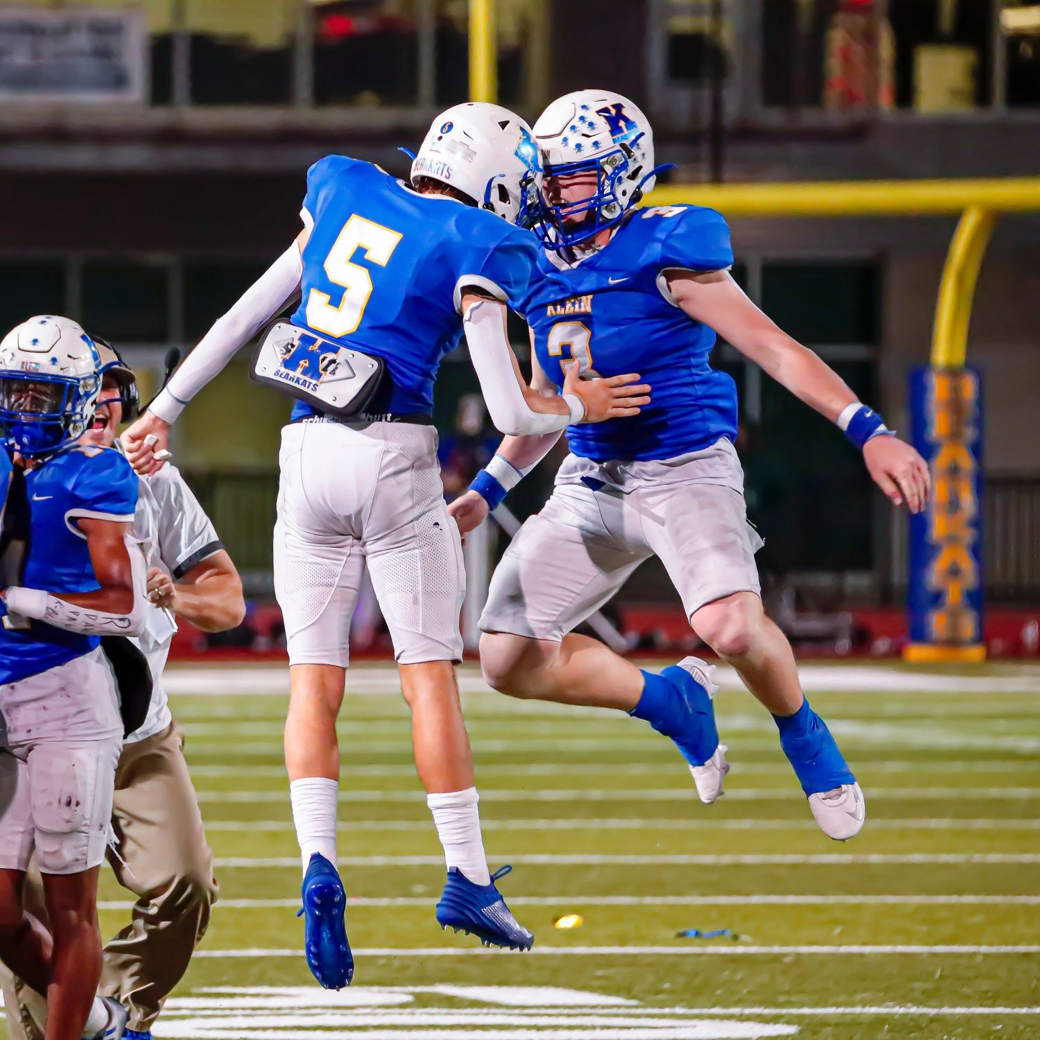 Two football players in blue and white uniforms celebrate with a mid-air jump bump during a game on the field.