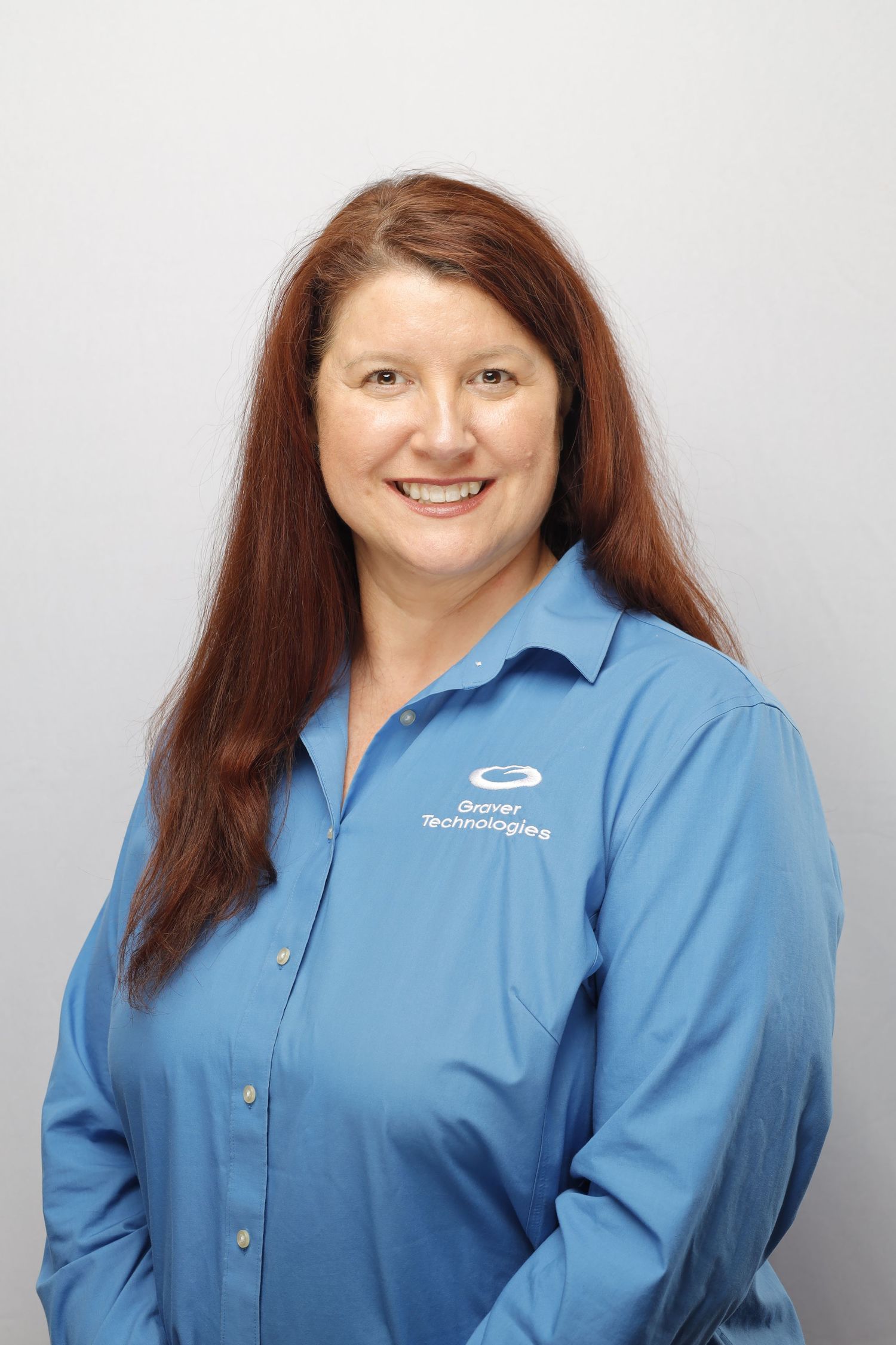 Professional headshot of a staff member in a light blue button-down company shirt with long auburn hair against a gray background.