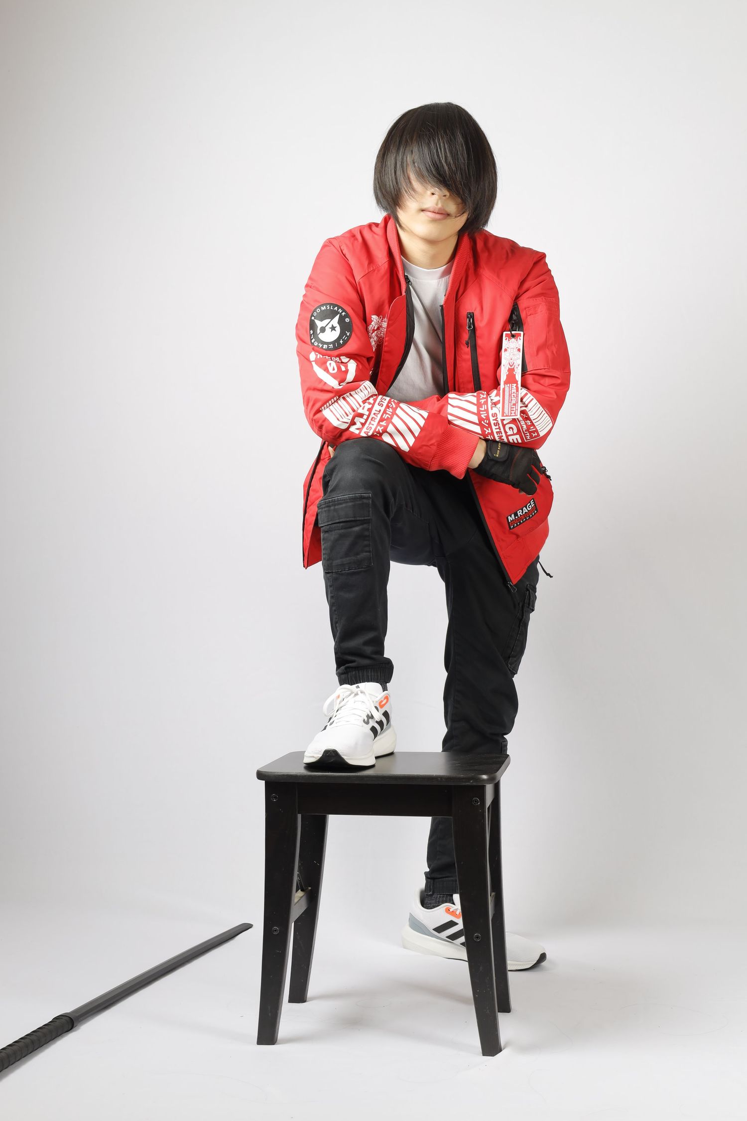 Person in red jacket and black pants posing on black stool against white background.
