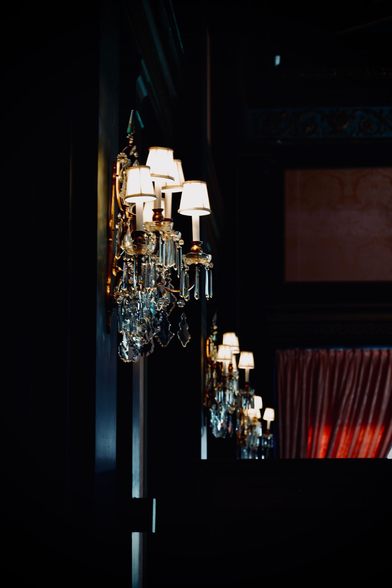 Elegant crystal chandelier with white lampshades illuminated in dark interior space.