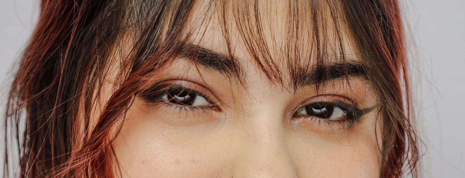 Close-up photo of eyes with warm brown irises and wispy bangs framing them against a light background.
