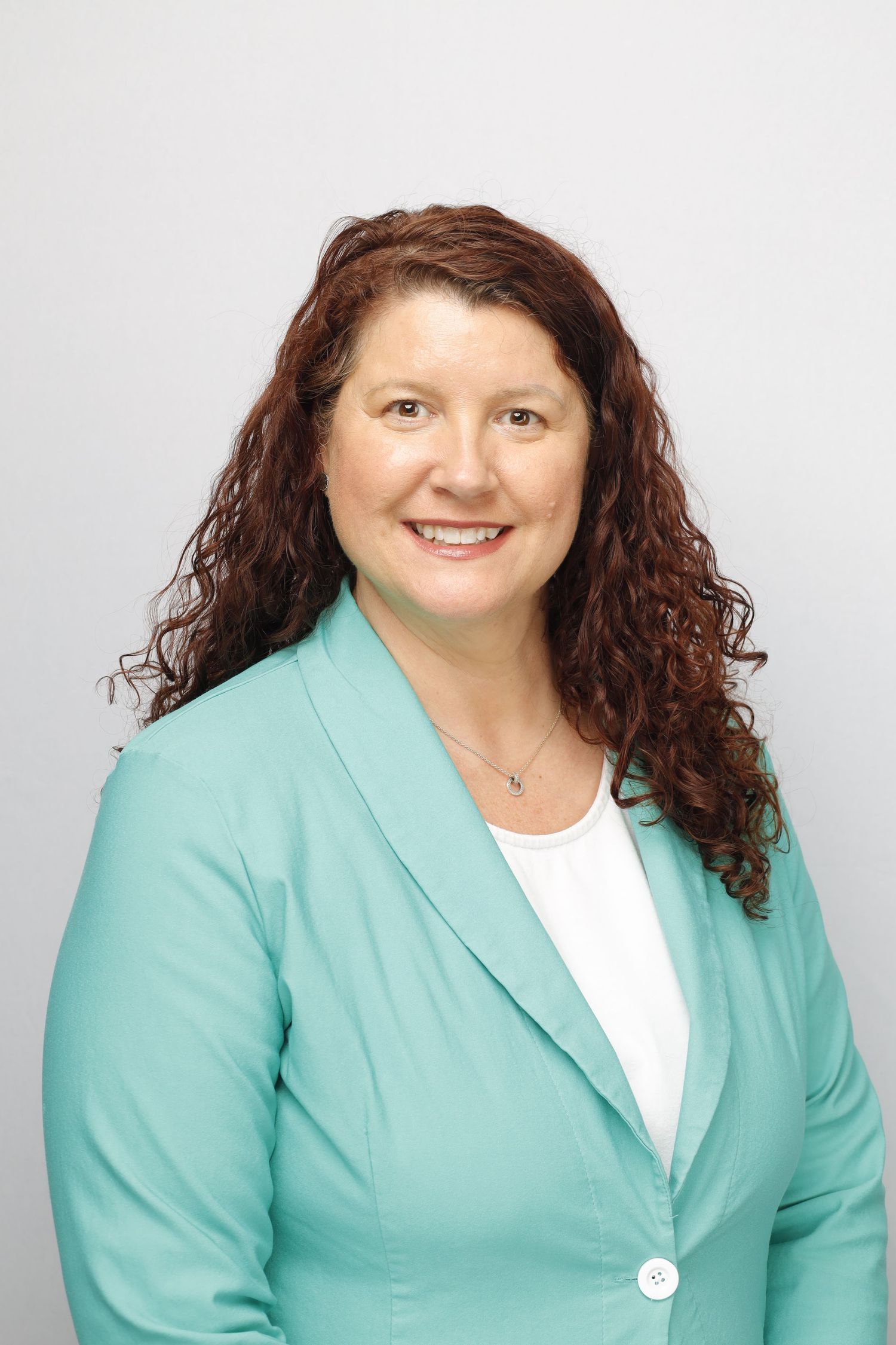Professional headshot of someone wearing a mint green blazer and white top with curly auburn hair against light background.
