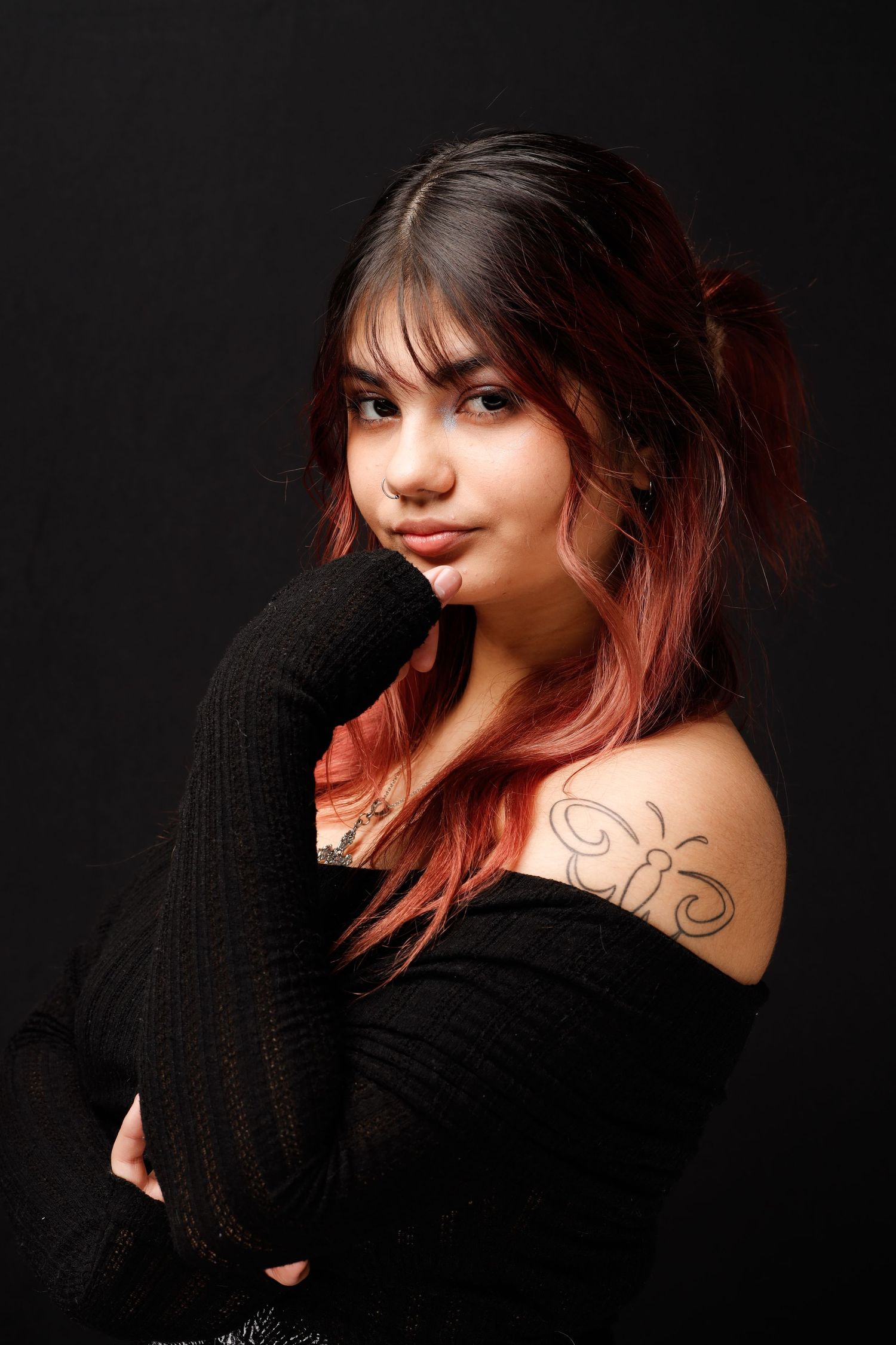Portrait of someone with red-tinted hair in a black sweater against dark background, showing a shoulder tattoo.