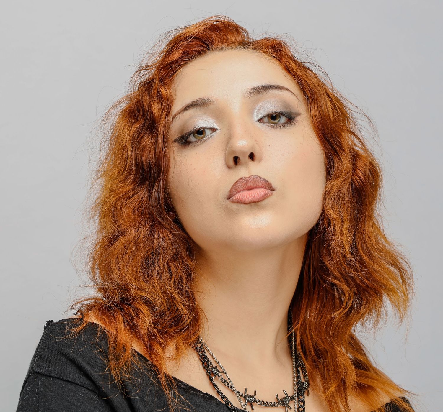 Person with vibrant copper red wavy hair wearing black leather jacket and silver necklace against grey background.