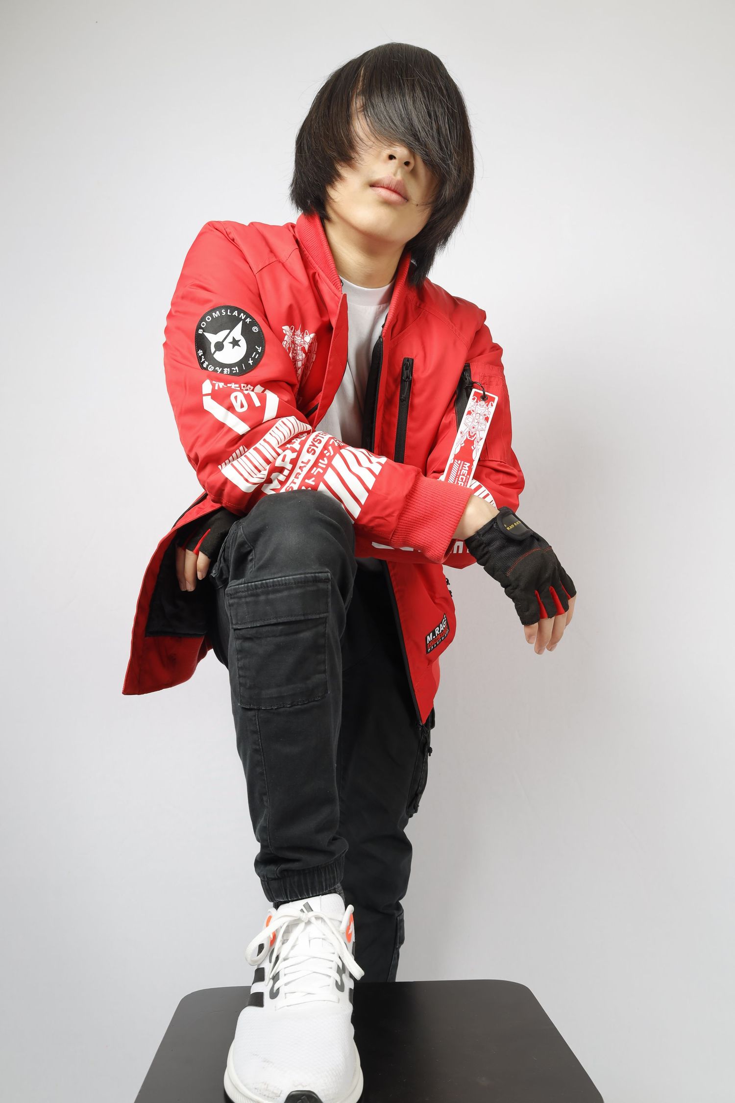 Person in red varsity jacket and black pants poses on platform wearing white sneakers in studio setting.