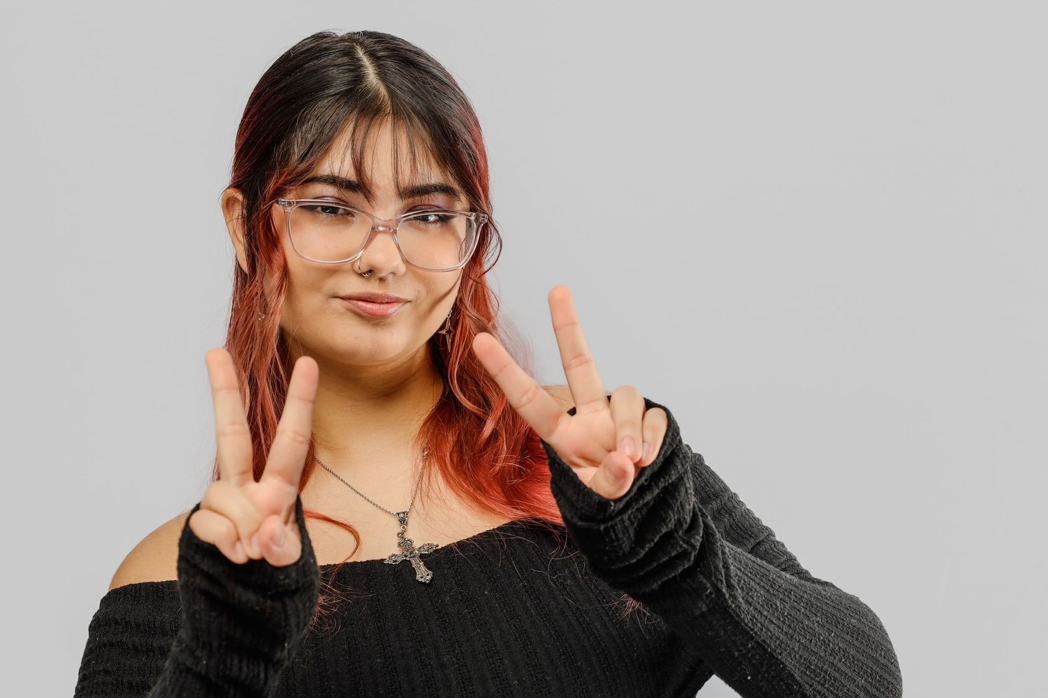 Someone with red-tinted hair wearing glasses and a black off-shoulder top makes peace signs against a gray background.