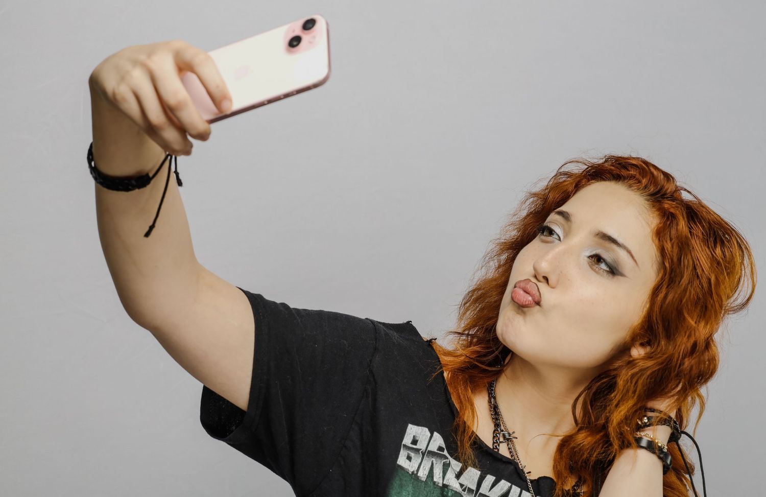 A person with red wavy hair makes a playful expression while taking a selfie on a smartphone against a gray background.