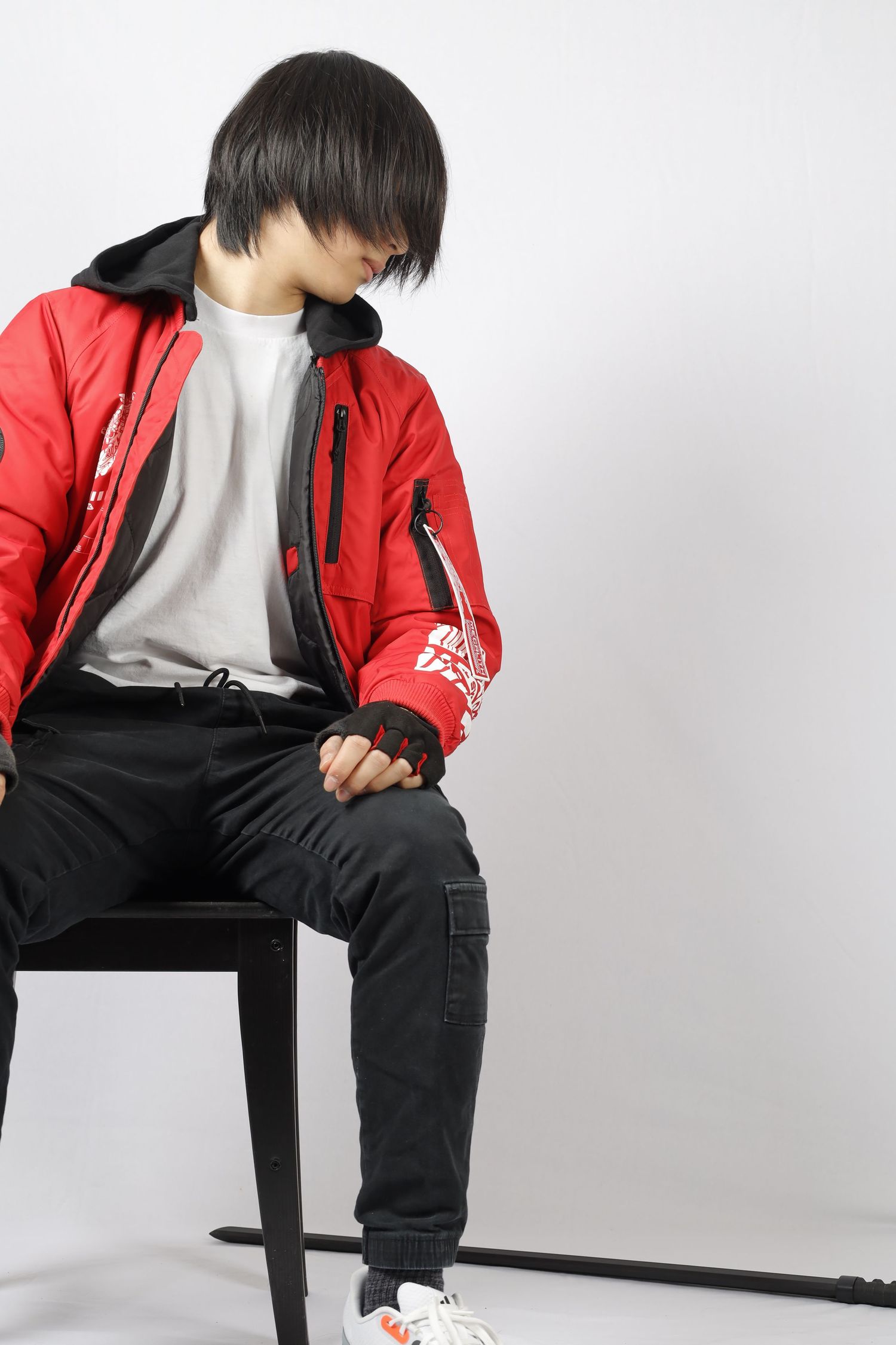 Person in red jacket and black pants sits on stool against white background wearing casual streetwear style clothing.