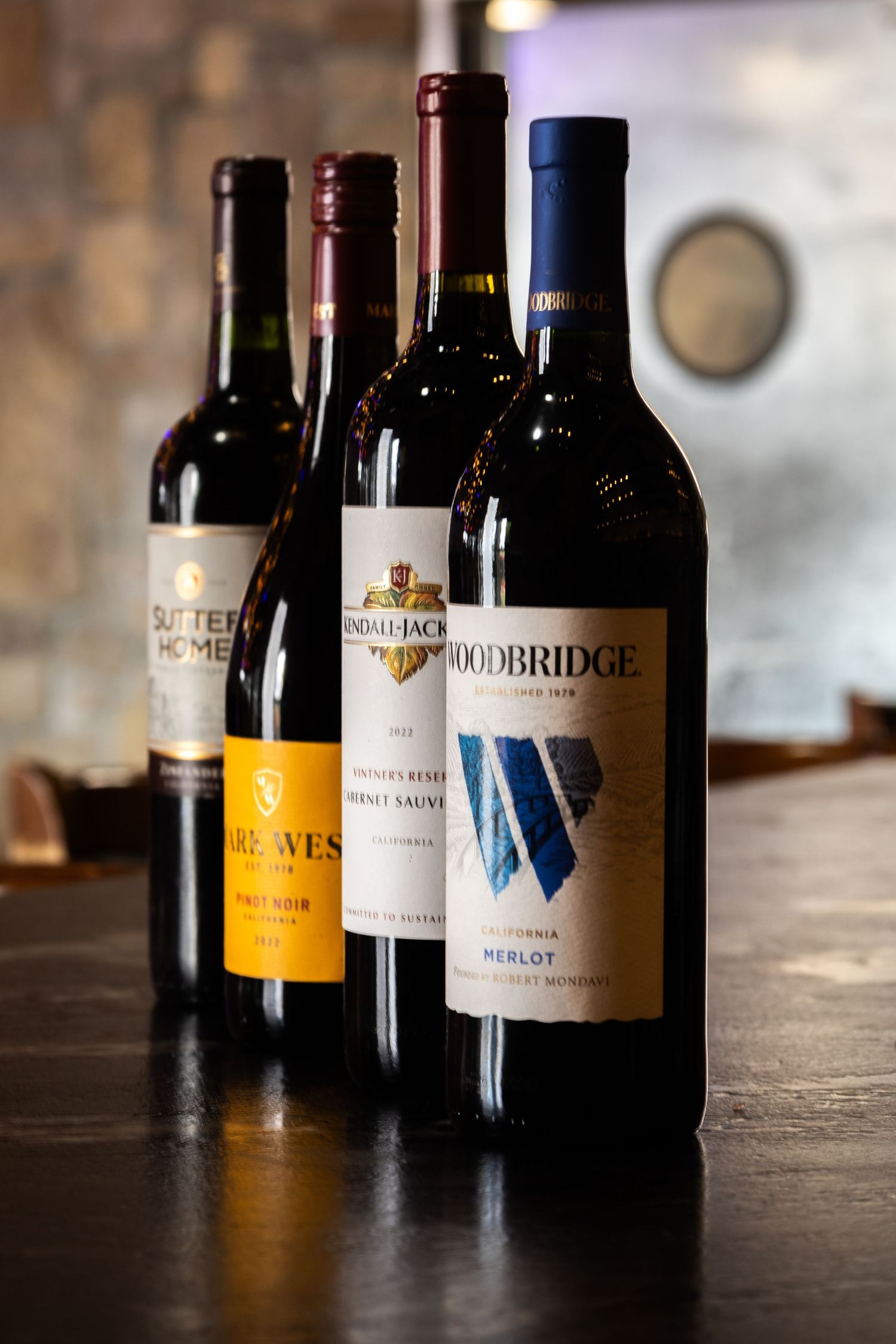 Four bottles of red wine including a Woodbridge Merlot stand in a row on a dark wooden surface with blurred background.