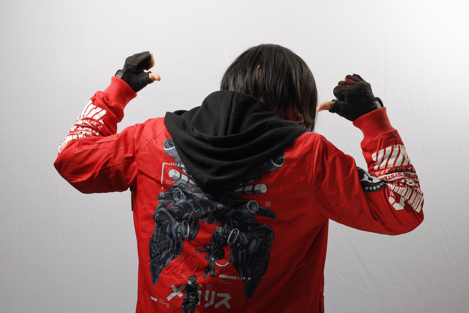 Figure in a red hoodie with graphic design detail seen from behind against a grey background raises both fists.