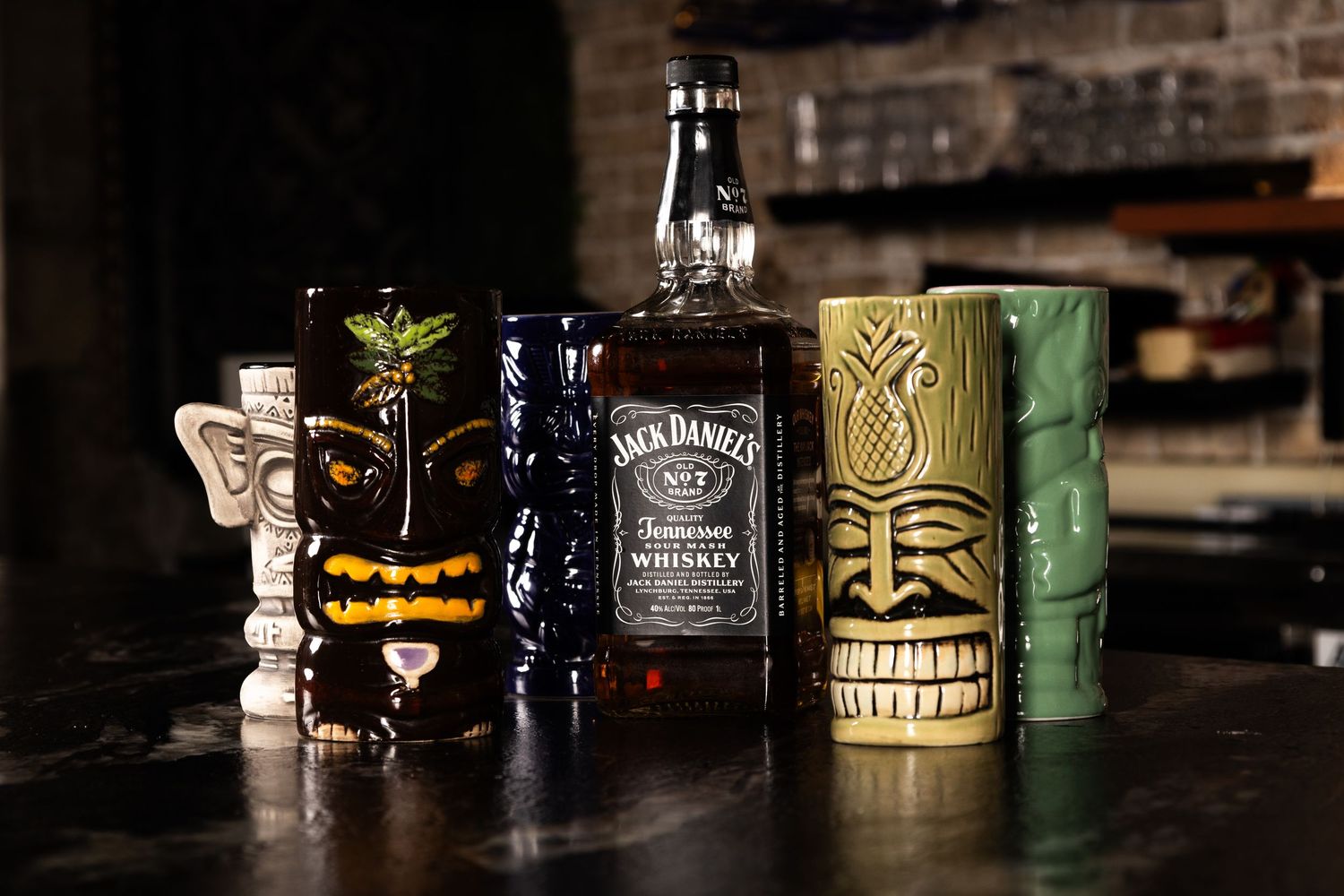 A collection of tiki mugs and a Jack Daniels whiskey bottle arranged on a dark wooden surface with mood lighting.