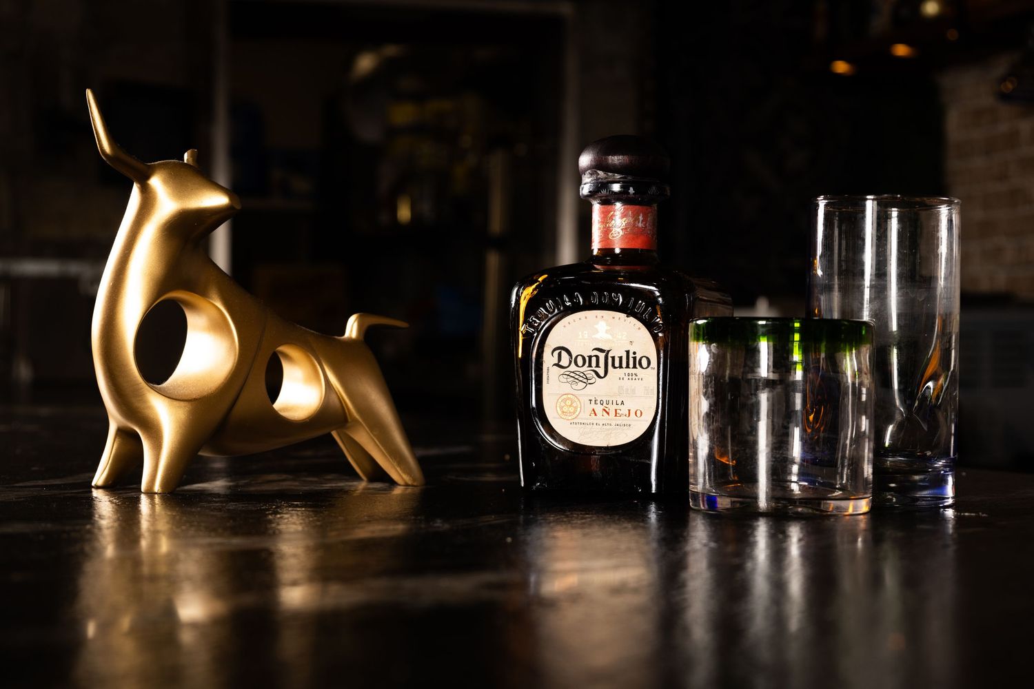 A stylish still life with Don Julio tequila bottle and glasses alongside a golden bull figurine in low light.