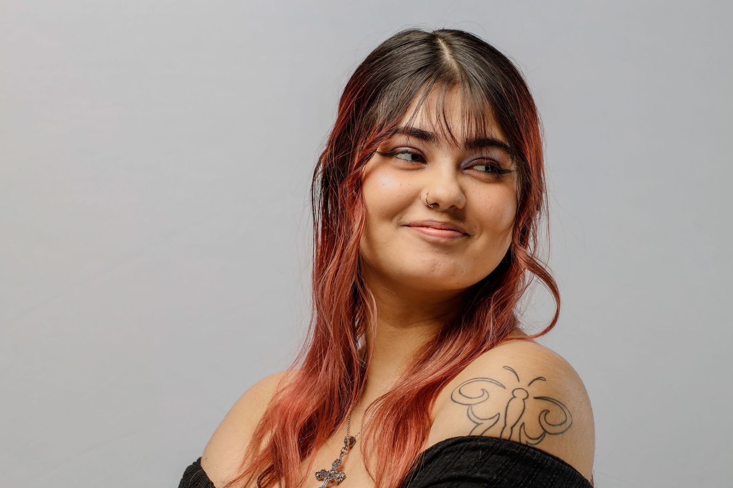 A woman with long pink-tinted hair and a visible arm tattoo smiles warmly against a gray background.
