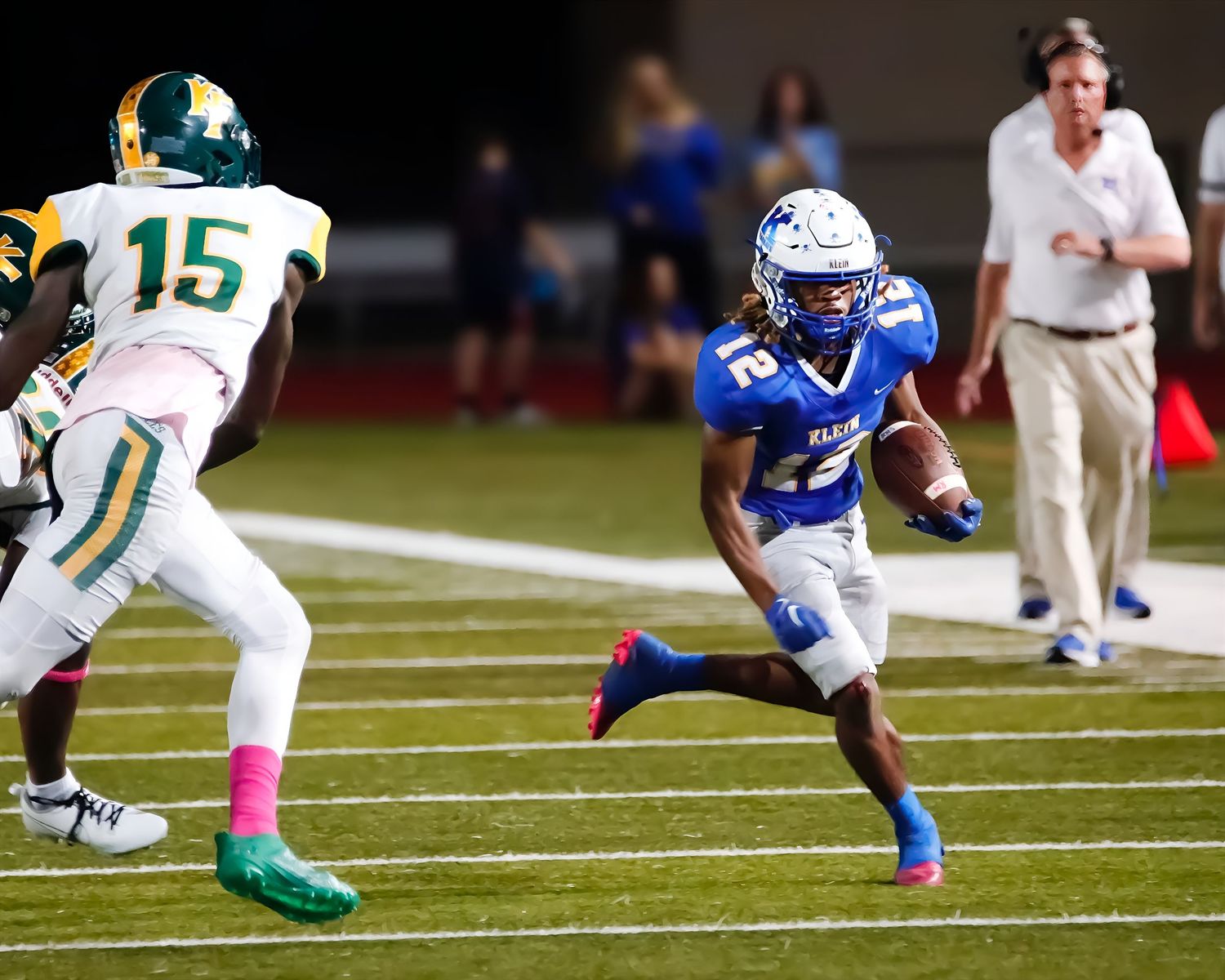 Two high school football players face off during a night game, with one in blue uniform and one in green and gold.