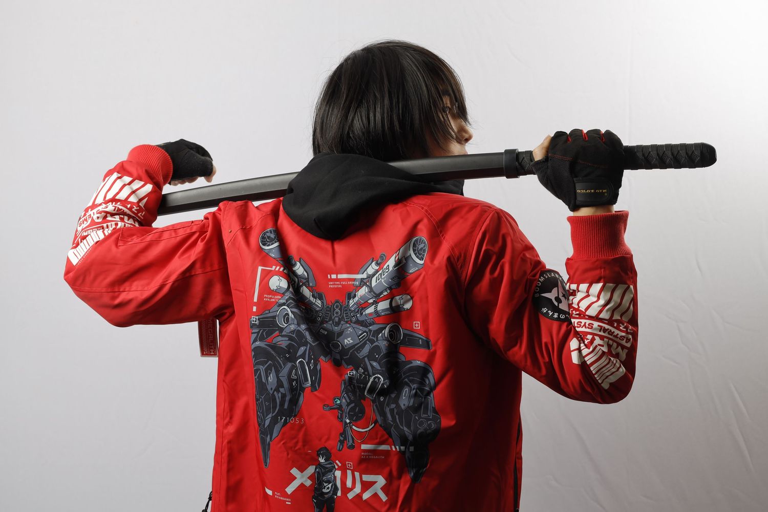 Person wearing red jacket with graphic design holds black pole or staff while facing away from camera against gray background.
