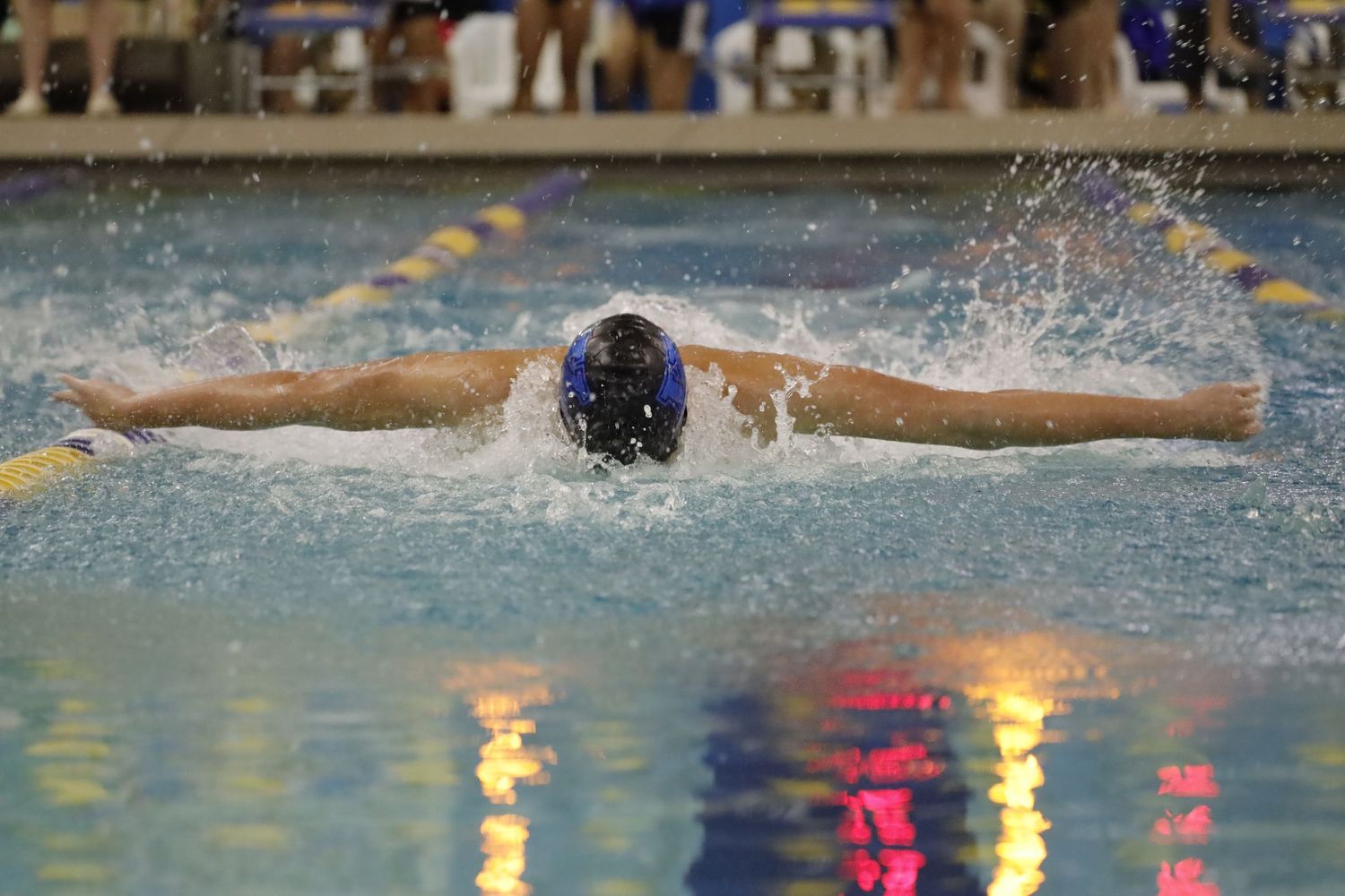 Competitive swimmer executes butterfly stroke during indoor swim meet with colorful reflections in pool water.