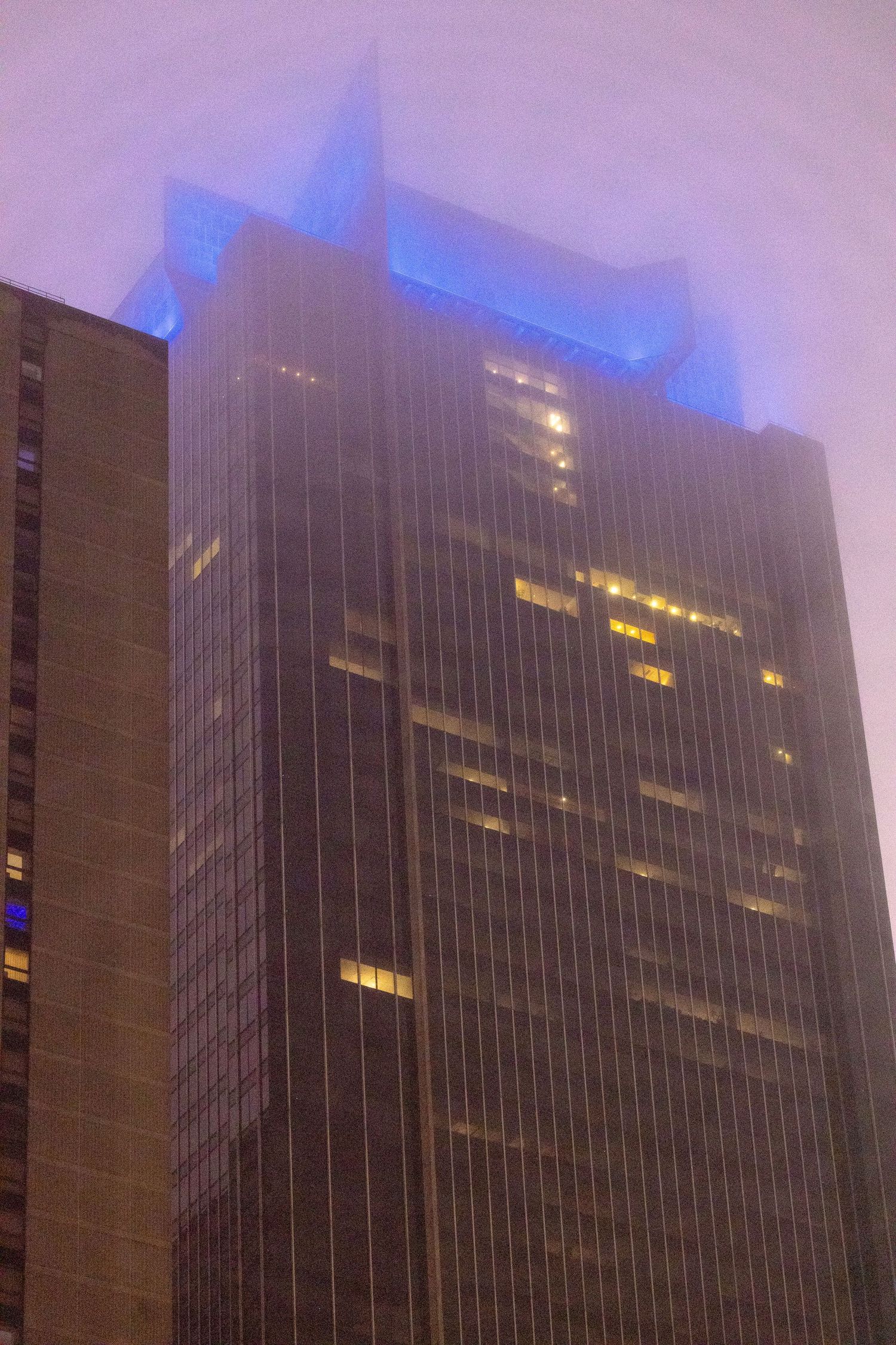 Modern skyscraper with illuminated blue crown glows against foggy purple sky at night.